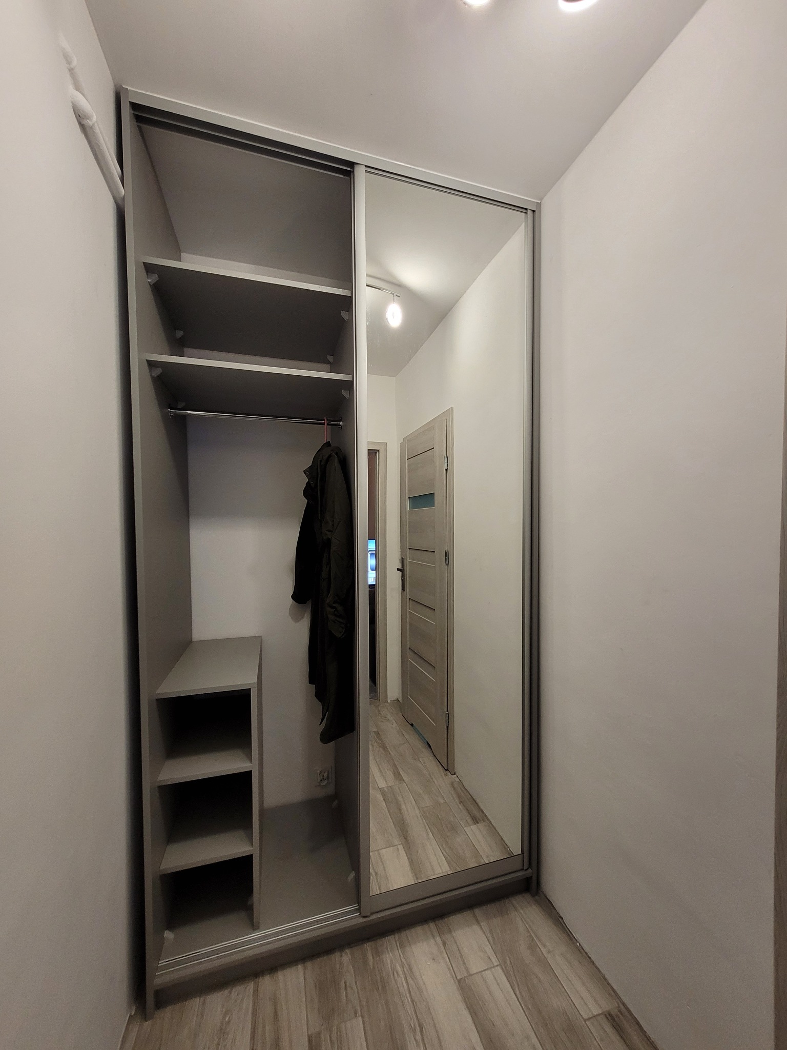 Szafa wnękowa z przesuwnymi drzwiami lustrzanymi, szara, z półkami i drążkiem na ubrania. Widok na wnętrze mieszkania w odbiciu lustra.
