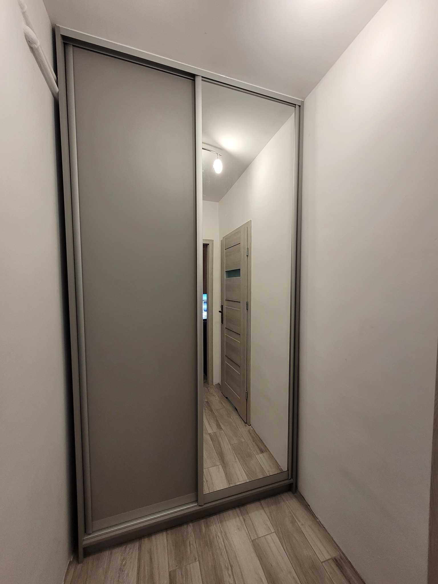 Szafa wnękowa z przesuwnymi drzwiami lustrzanymi, szara, wkomponowana w korytarz. Widoczne odbicie wnętrza mieszkania w lustrze.