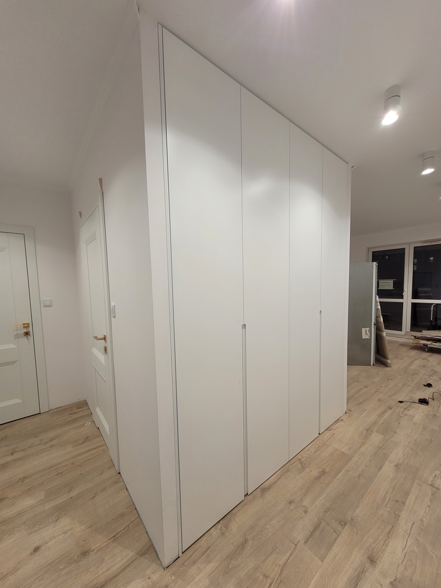 Biała szafa wnękowa z minimalistycznymi uchwytami, dopasowana do wysokości pomieszczenia, na tle drewnianej podłogi i białych drzwi.