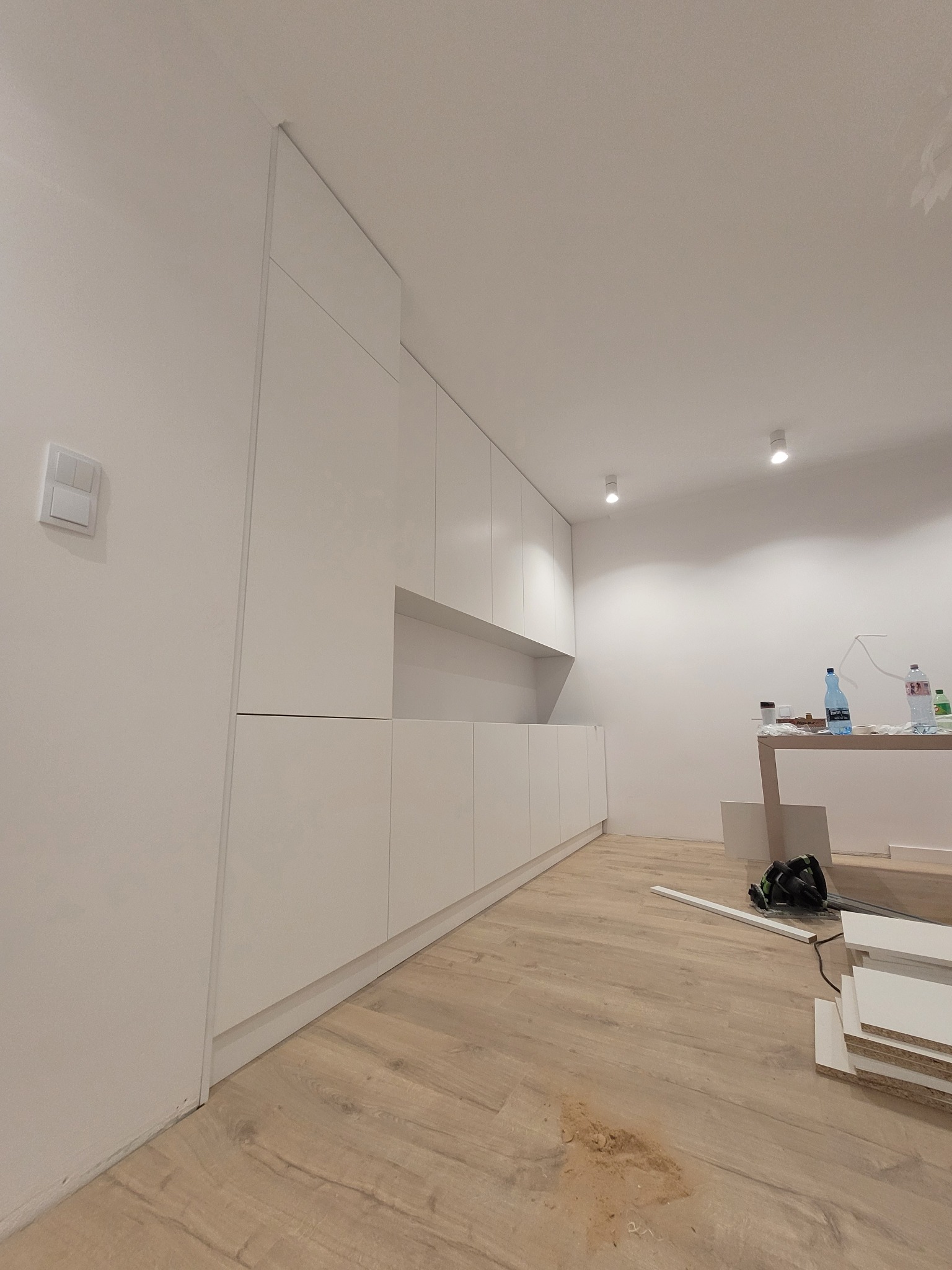 Minimalistyczna, biała zabudowa kuchenna z długim blatem, widoczna w trakcie montażu. Na podłodze leżą narzędzia i elementy wykończeniowe. Oświetlenie sufitowe punktowe.