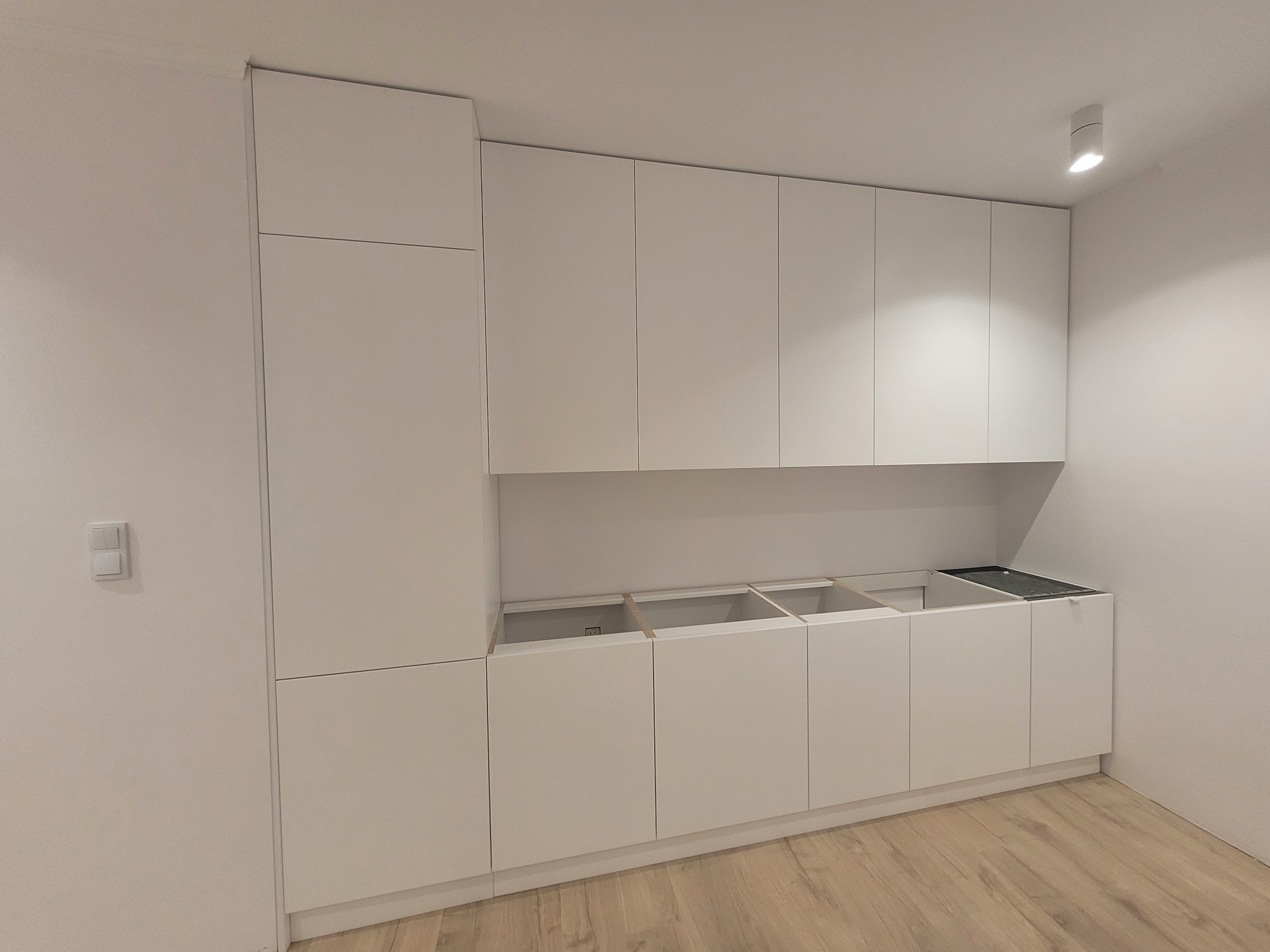 Minimalistyczna, biała zabudowa kuchenna w trakcie montażu, widoczne puste przestrzenie pod blatem, oświetlenie punktowe, jasna podłoga. Nowoczesny design.