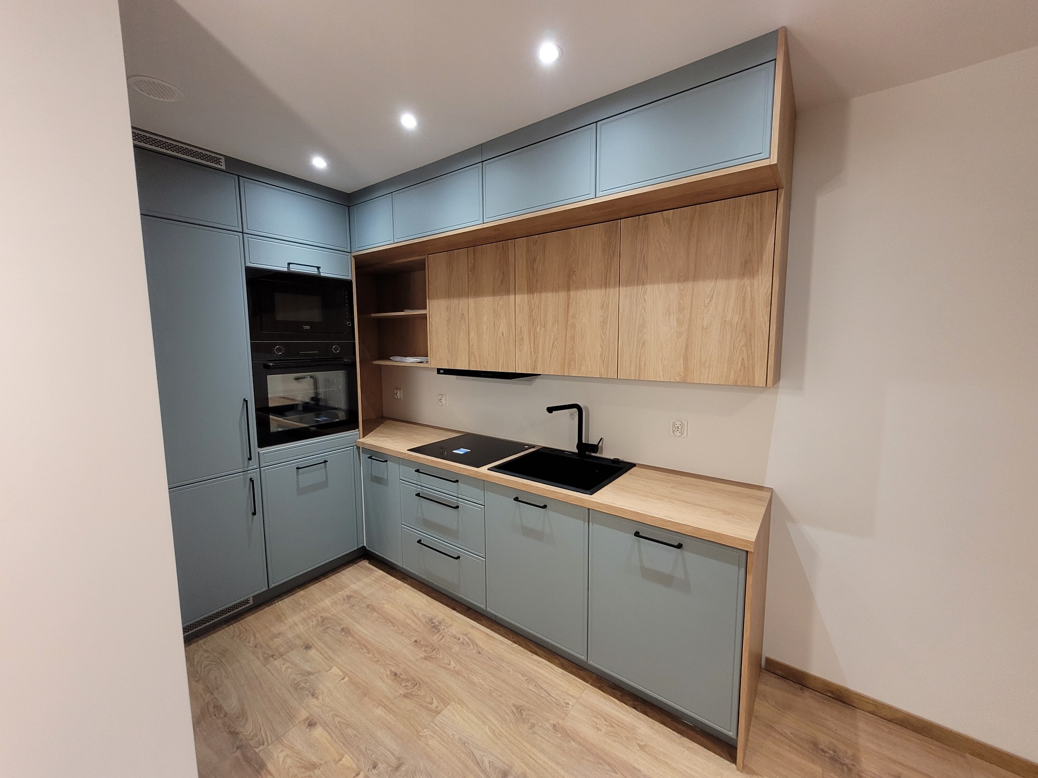 Narożna kuchnia z błękitnymi szafkami i drewnianymi blatami. Nowoczesny design z czarną armaturą i sprzętem AGD. Jasne drewno na podłodze.