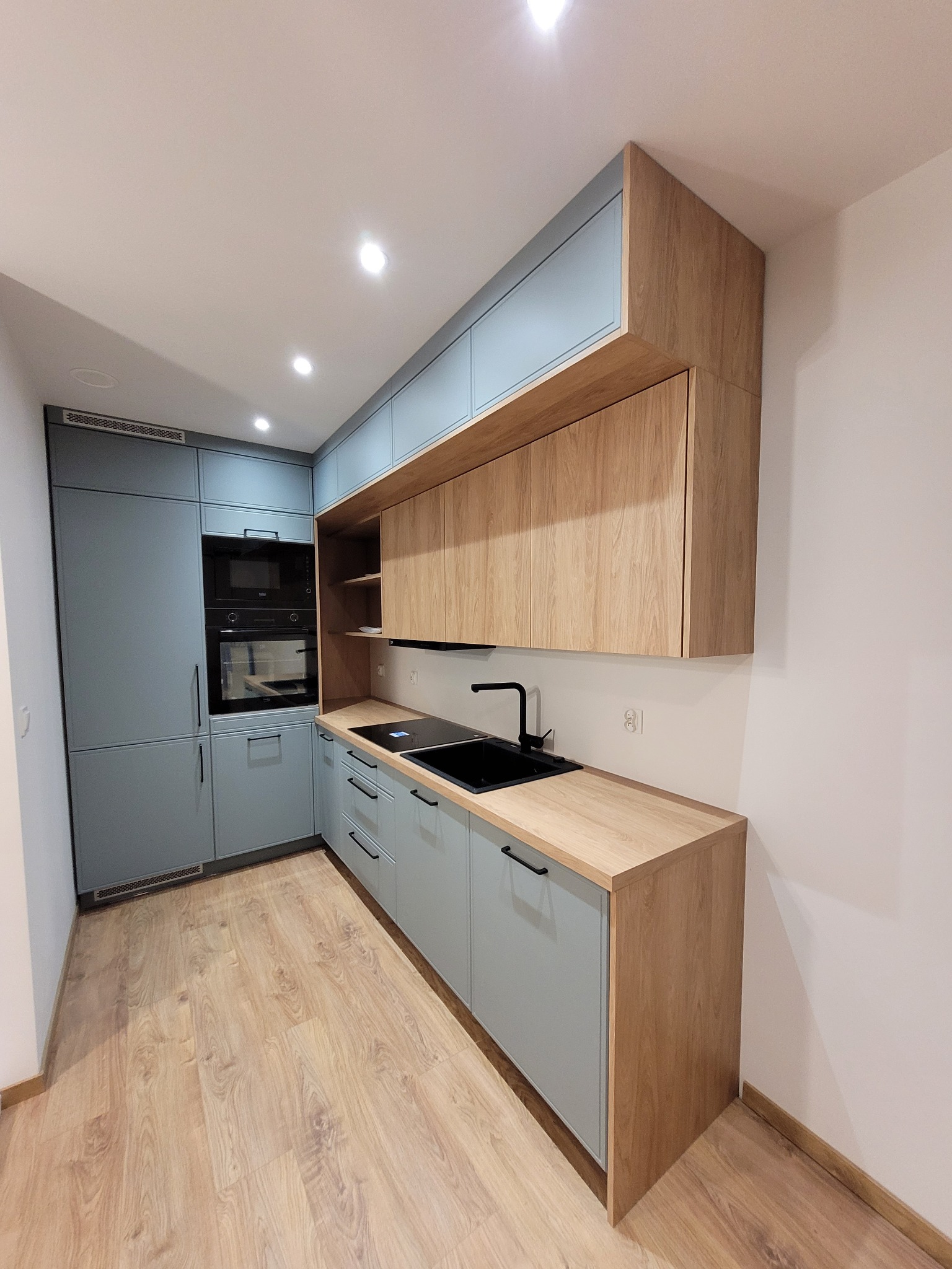 Nowoczesna kuchnia z błękitnymi frontami i drewnianymi blatami. Czarny zlew i bateria, zabudowane sprzęty AGD. Jasna podłoga i oświetlenie punktowe.