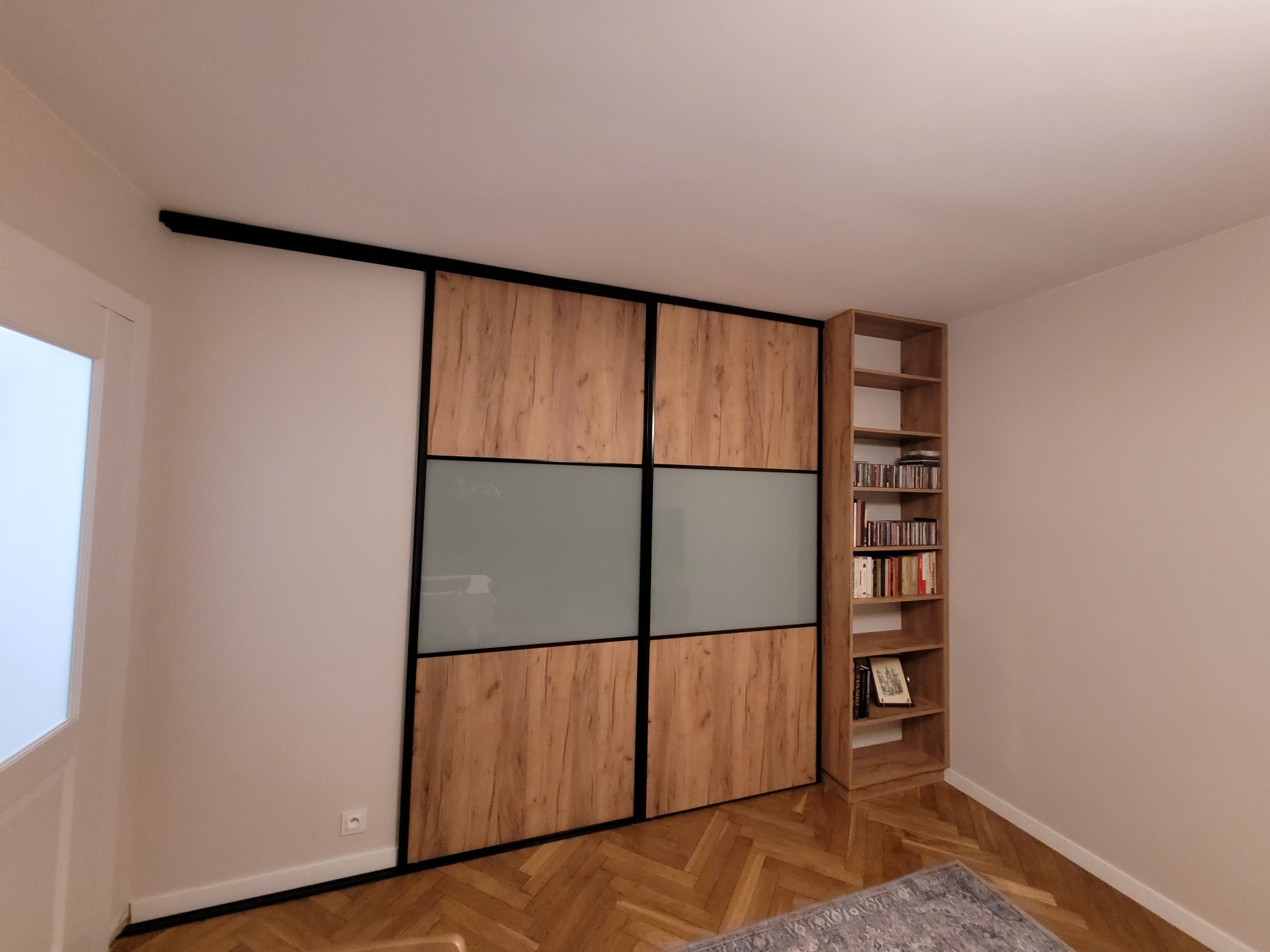 Szafa wnękowa z przesuwnymi drzwiami, drewnopodobne panele i matowe szkło, czarne obramowanie, obok regał na książki, parkiet w jodełkę. Nowoczesny design.