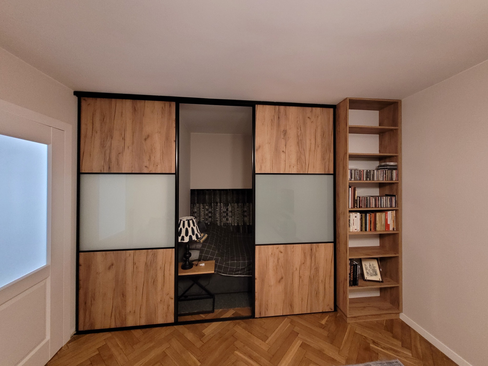 Szafa wnękowa z przesuwnymi drzwiami z drewnopodobną okleiną i mlecznym szkłem, wbudowana w ścianę, obok regał na książki z tej samej okleiny.