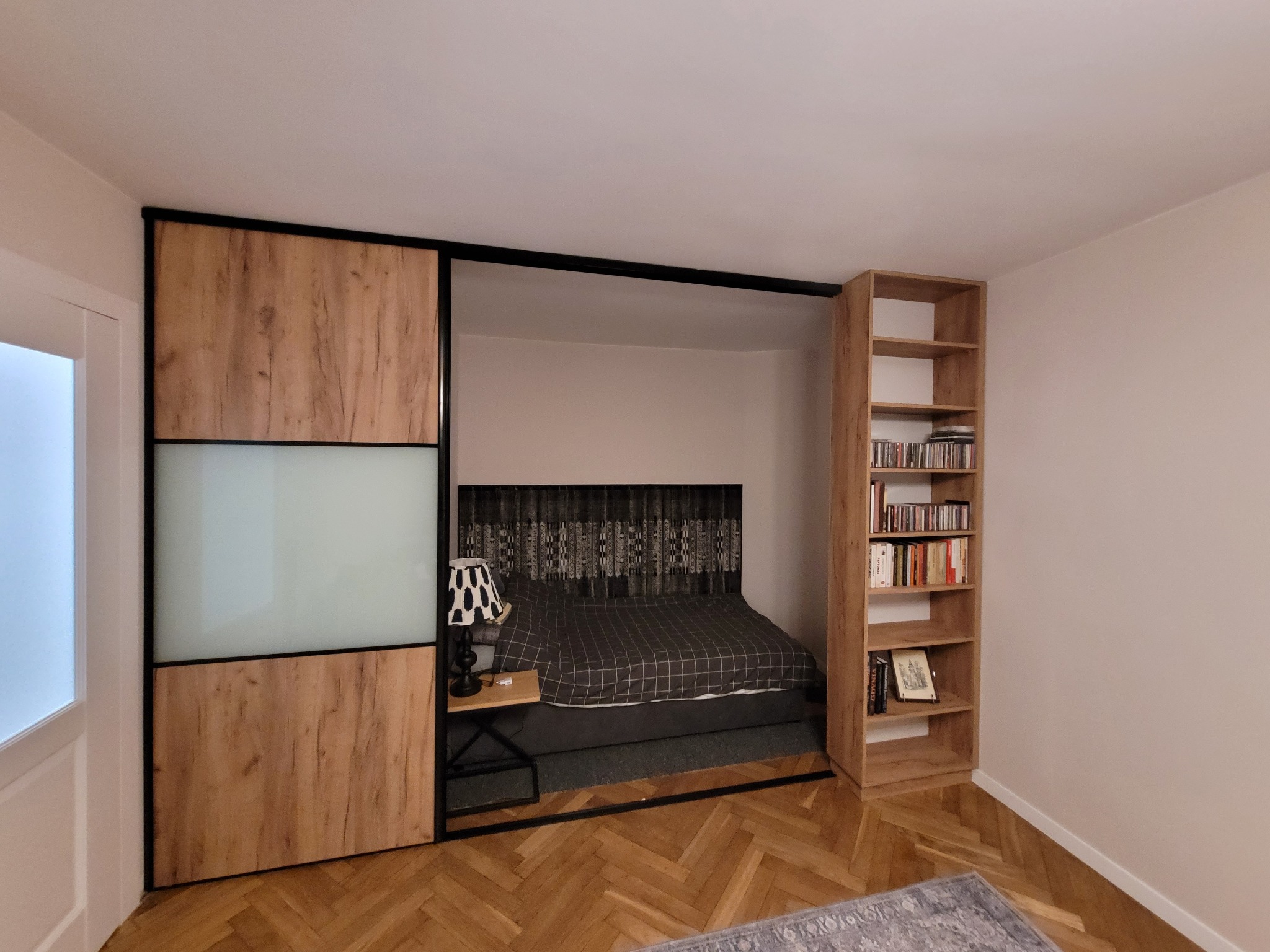 Nowoczesna szafa przesuwna z drewnianymi panelami i mlecznym szkłem, wkomponowana w aranżację sypialni z łóżkiem i regałem na książki. Parkiet w jodełkę.