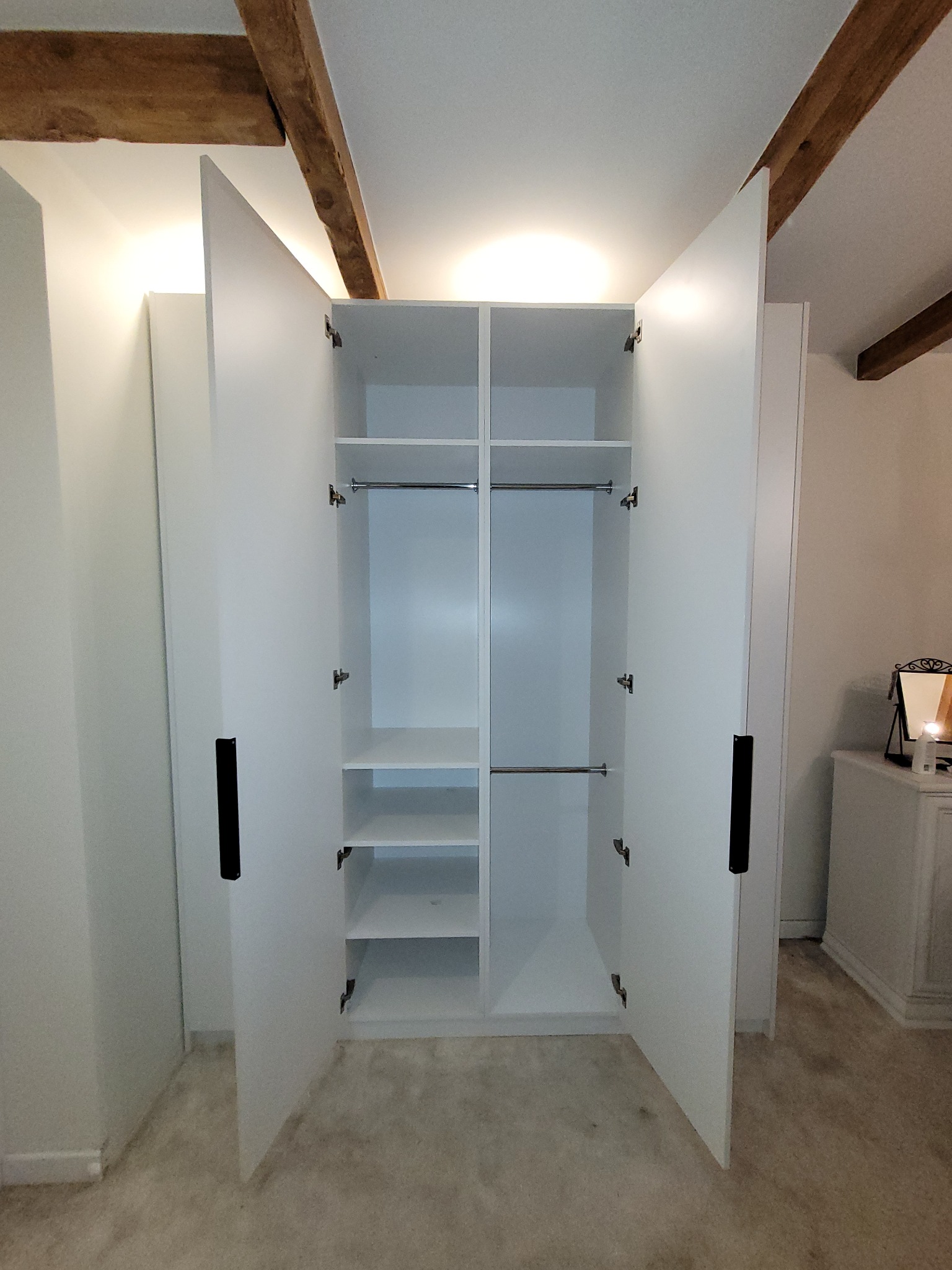 Biała szafa wnękowa z otwartymi drzwiami, widoczne półki i drążki na ubrania. Uwagę przykuwają drewniane belki stropowe w tle.