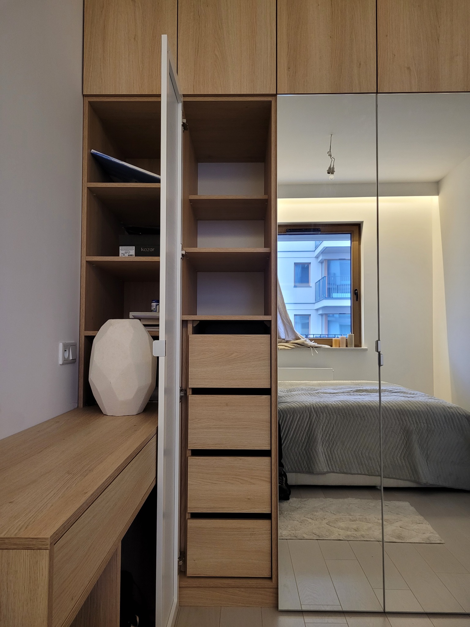 Jasna szafa z lustrzanymi drzwiami, wbudowana w pokój. Widoczne półki, szuflady i biurko. W lustrze odbija się łóżko i okno z widokiem na budynek.