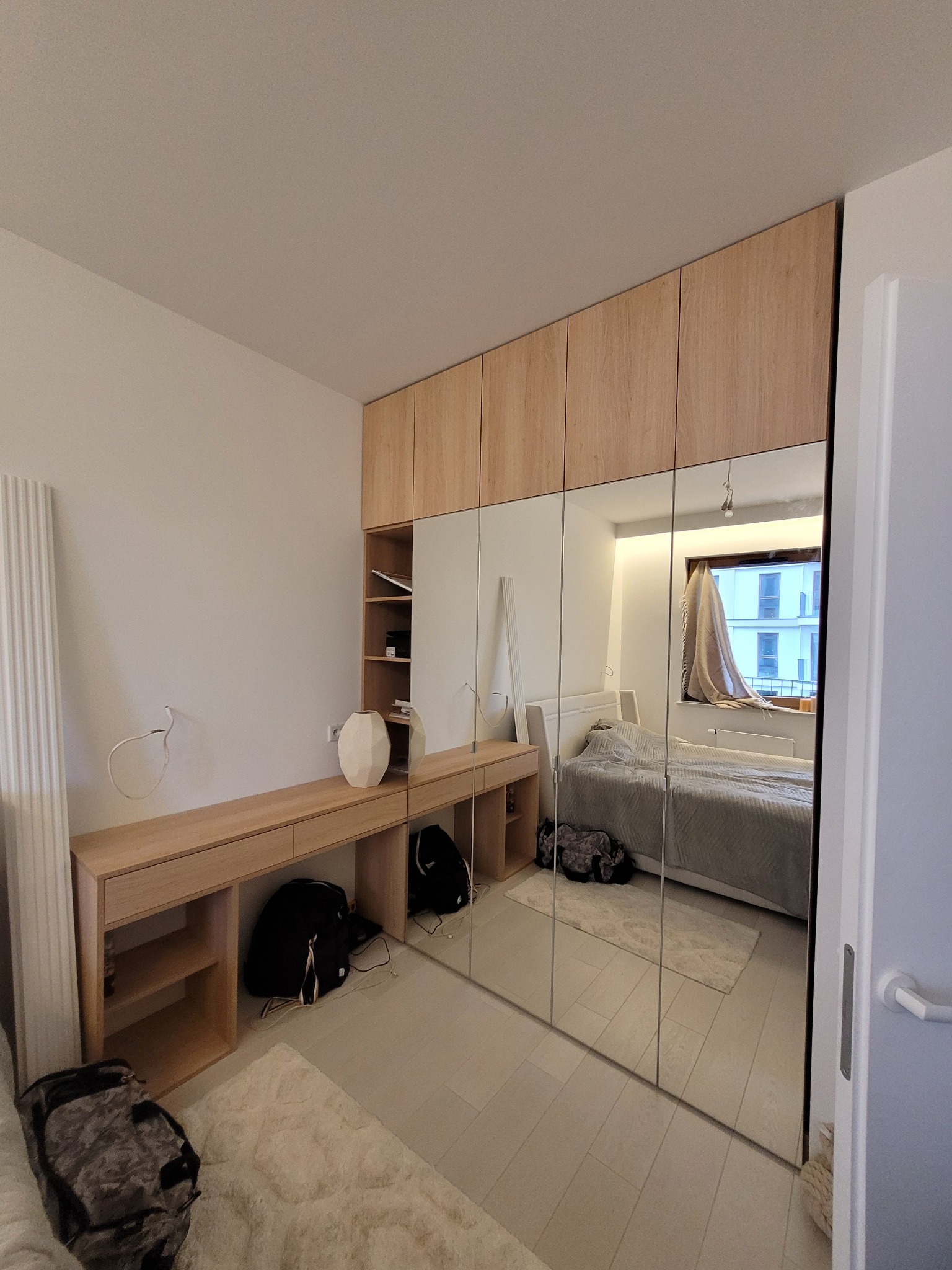 Jasna szafa wnękowa z lustrzanymi drzwiami, zintegrowana z toaletką i półkami, w nowoczesnej sypialni. Widoczne odbicie łóżka i okna.