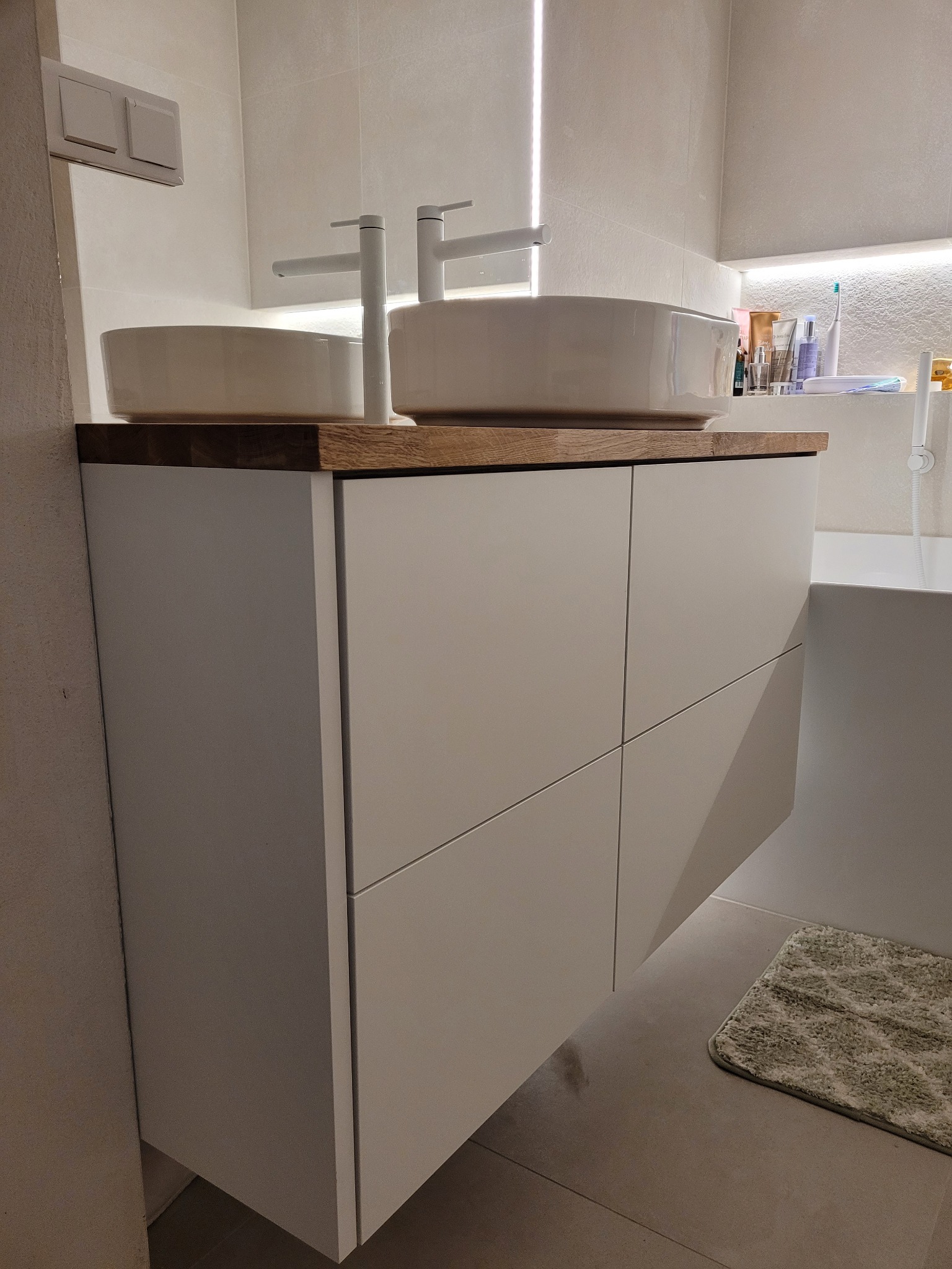 Nowoczesna łazienka: biała szafka pod umywalkę z drewnianym blatem, dwie okrągłe umywalki i minimalistyczne baterie. W tle oświetlona półka z kosmetykami.