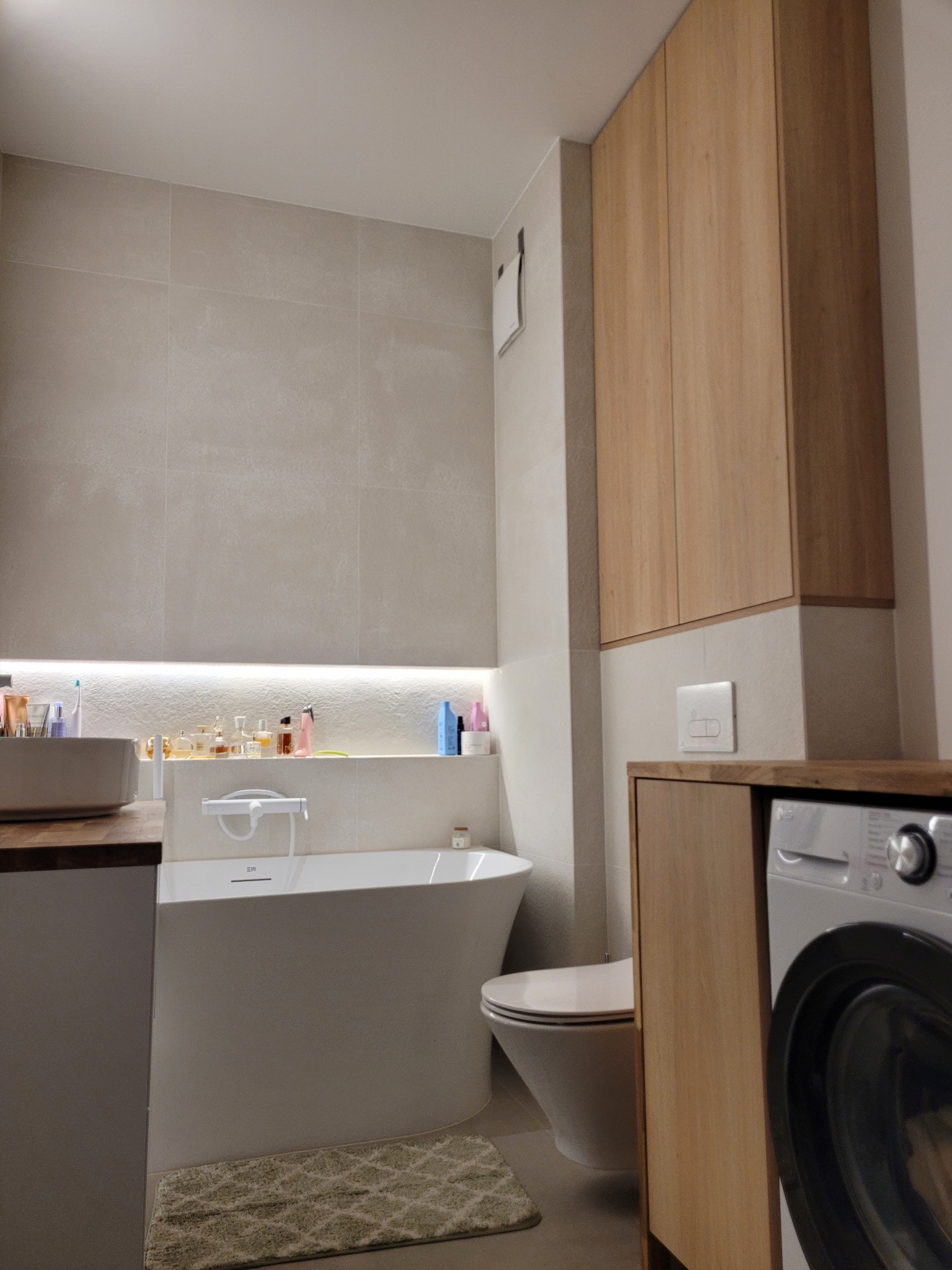 Nowoczesna łazienka z drewnianymi szafkami, wanną i pralką. Minimalistyczny design, neutralna kolorystyka, oświetlenie LED. Umywalka z drewnianym blatem.