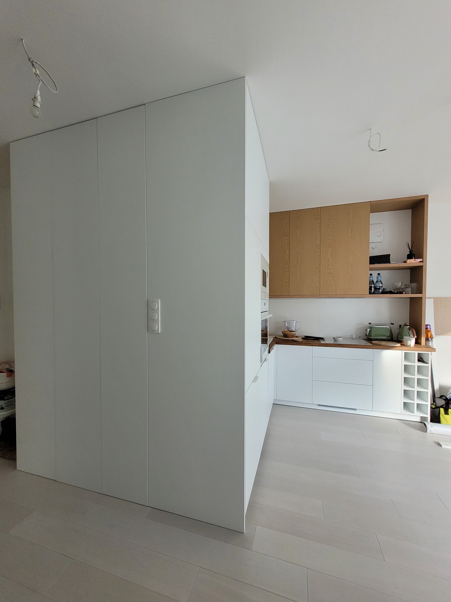 Nowoczesna, minimalistyczna kuchnia z zabudową szaf w kolorze białym i drewnianymi elementami. Widoczna zabudowa od podłogi do sufitu z dyskretnymi uchwytami.