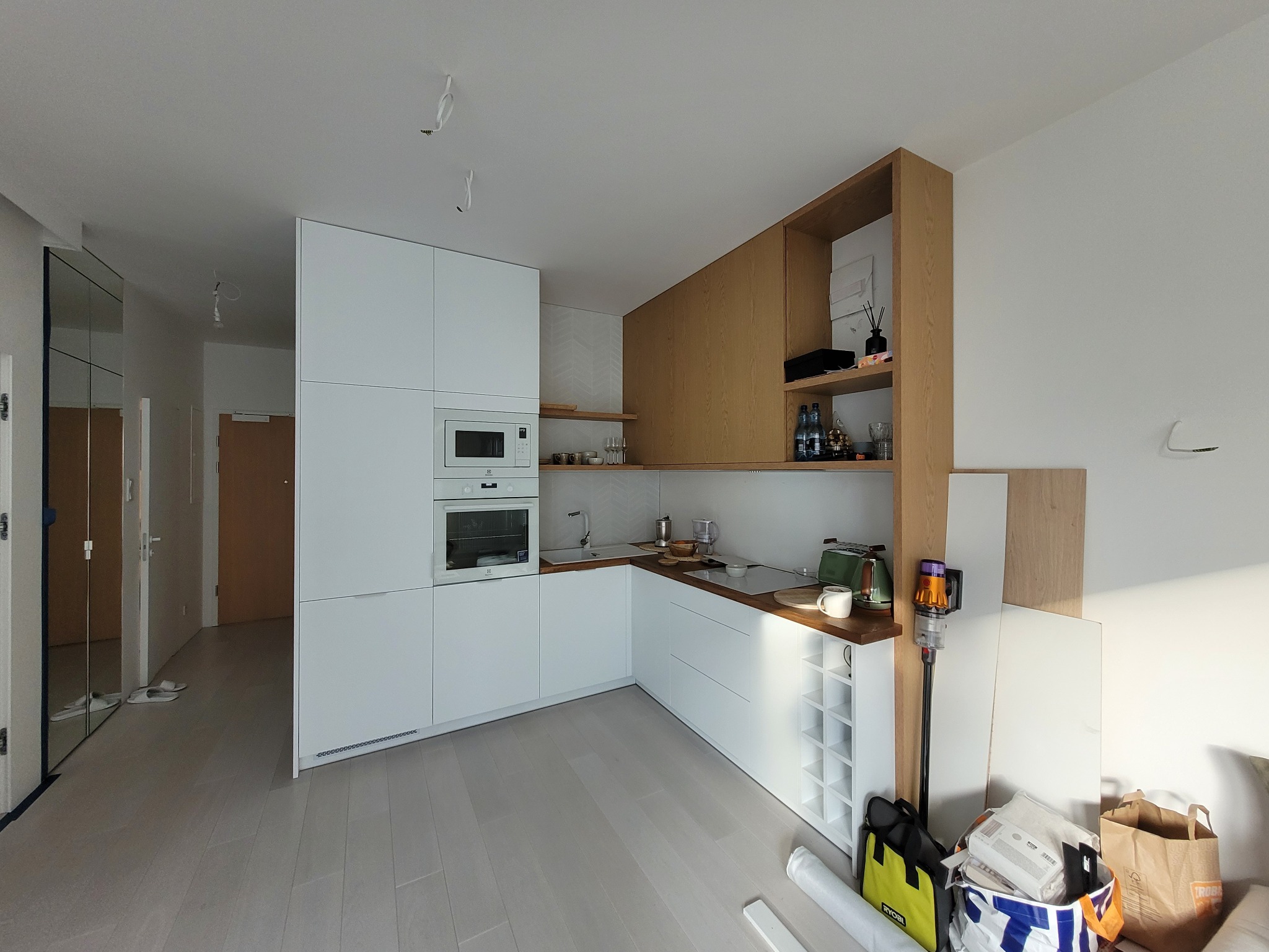 Nowoczesna, jasna kuchnia z białymi frontami i drewnianymi akcentami. Widoczne sprzęty AGD w zabudowie i elementy wyposażenia. Częściowo widoczne torby i akcesoria montażowe.
