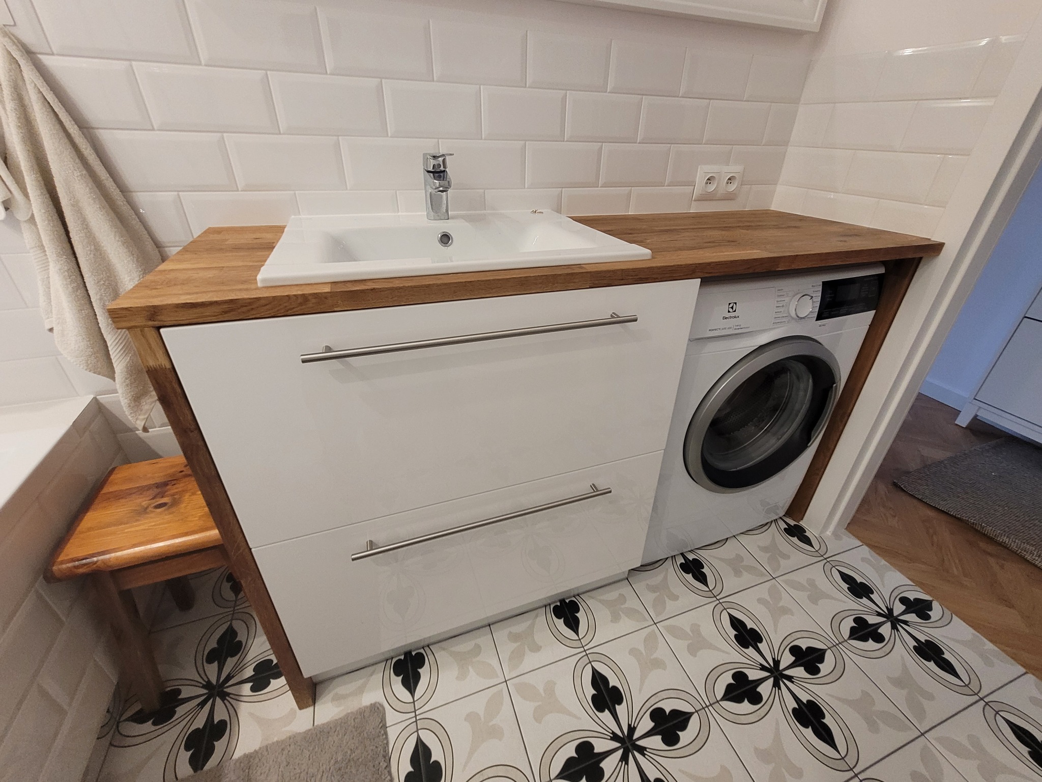 Biała szafka łazienkowa z drewnianym blatem, wbudowana pralka Electrolux. Umywalka nablatowa, bateria chromowana. Dekoracyjne płytki podłogowe.