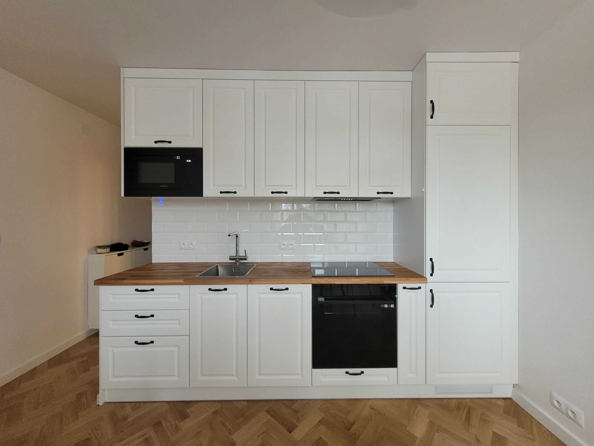 Biała kuchnia na wymiar z drewnianym blatem i czarnymi uchwytami, widok z przodu, parkiet w jodełkę na podłodze, nowoczesny styl.