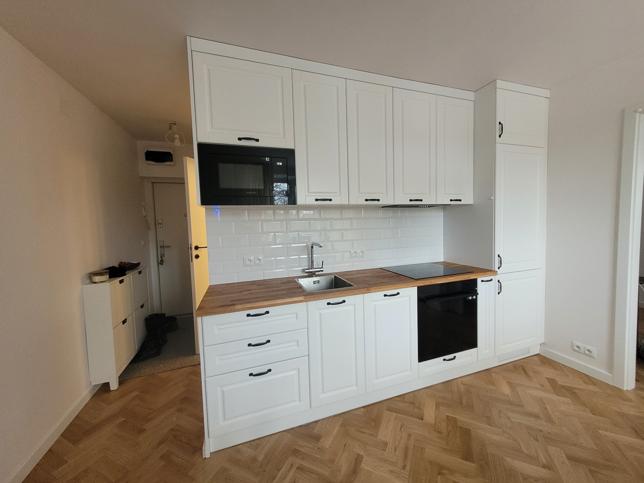 Biała zabudowa kuchenna z drewnianym blatem i czarnymi uchwytami, wkomponowana w jasne wnętrze z parkietem w jodełkę. Widoczny sprzęt AGD.