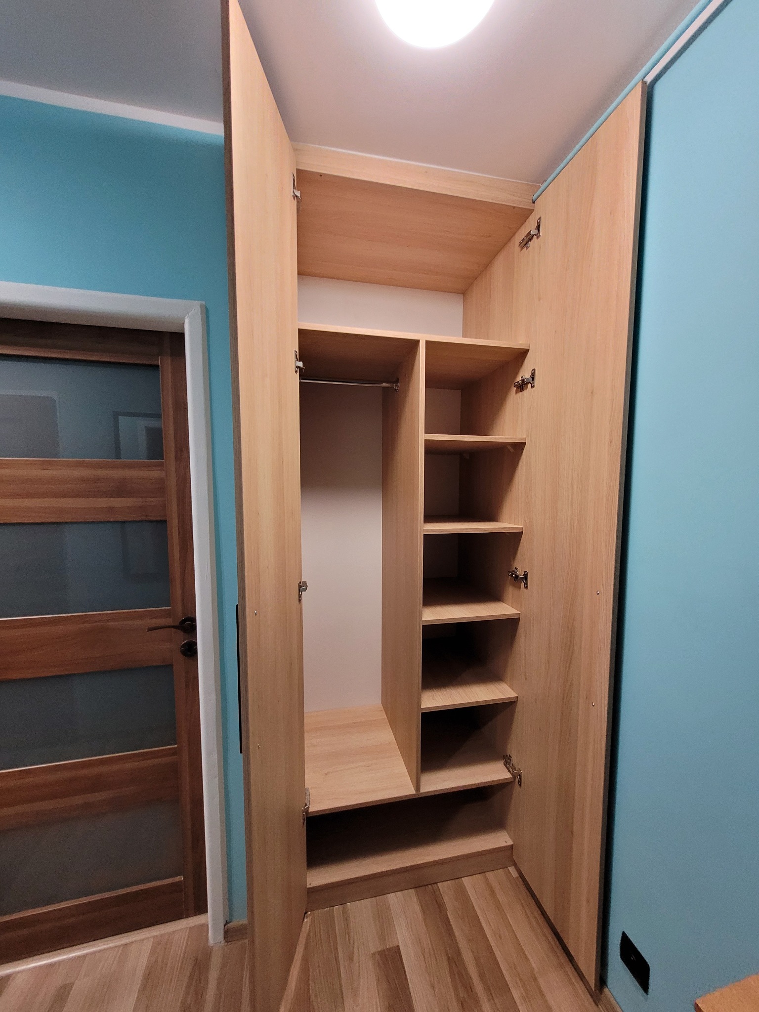 Jasna, drewniana szafa z otwartymi drzwiami, ukazująca półki i drążek na ubrania. Widoczna drewniana podłoga i drzwi w tle, na tle turkusowej ściany.