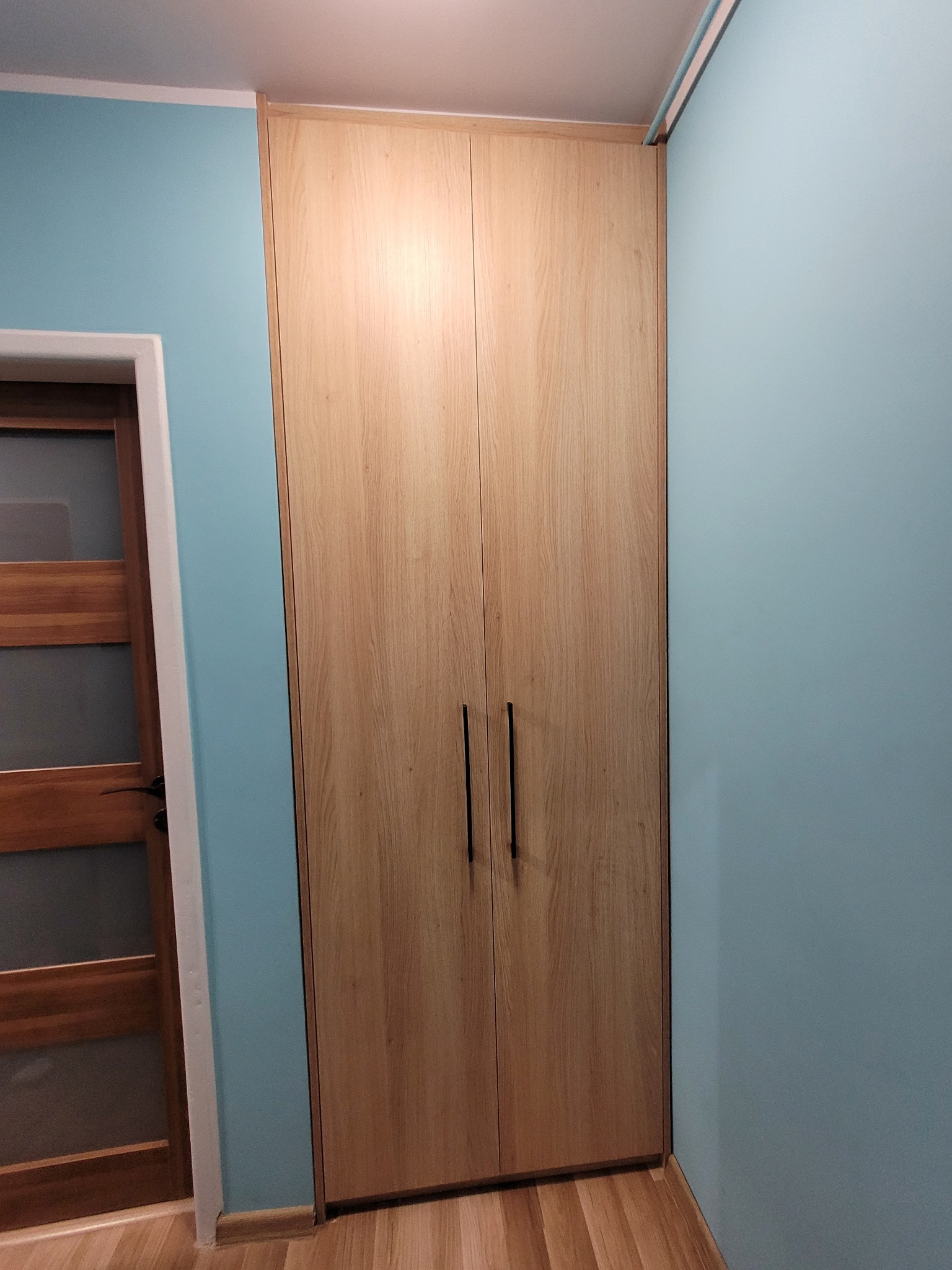Jasna, drewniana szafa dwudrzwiowa z długimi, czarnymi uchwytami, wkomponowana we wnękę pokoju z błękitnymi ścianami i drewnianą podłogą. Minimalistyczny design.
