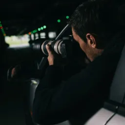 Detektyw w czarnej bluzie, zza szyby samochodu, robi zdjęcie profesjonalnym aparatem z teleobiektywem w ciemnym garażu z rzędami zielonych świateł w tle.