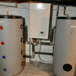 Dwa szare zbiorniki akumulacyjne EMEI po bokach białego kotła YUTAKI z wyświetlaczem, rury instalacji grzewczej z izolacją termiczną na tle białych płytek.