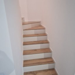 Ekipa Alana - Bieg schodów w jasnej klatce schodowej, wykończony panelami imitującymi drewno, z białymi podstopnicami, widziany z dołu.