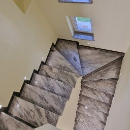 Ekipa Alana - Schody wykończone marmurem z podświetleniem LED, widok z góry, z fragmentem drewnianej podłogi i butami. Dwa okna dachowe w tle.