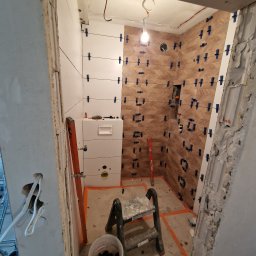 Ekipa Alana - Montaż płytek na ścianie w łazience, widoczne płytki w dwóch kolorach: białe i brązowe ułożone w jodełkę, niebieskie krzyżyki dystansowe, poziomica i drabinka malarska.