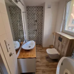 Nowoczesna łazienka z owalną umywalką na drewnianym blacie, mozaikową ścianą pod prysznicem i podwieszaną toaletą, wykończona płytkami imitującymi drewno.