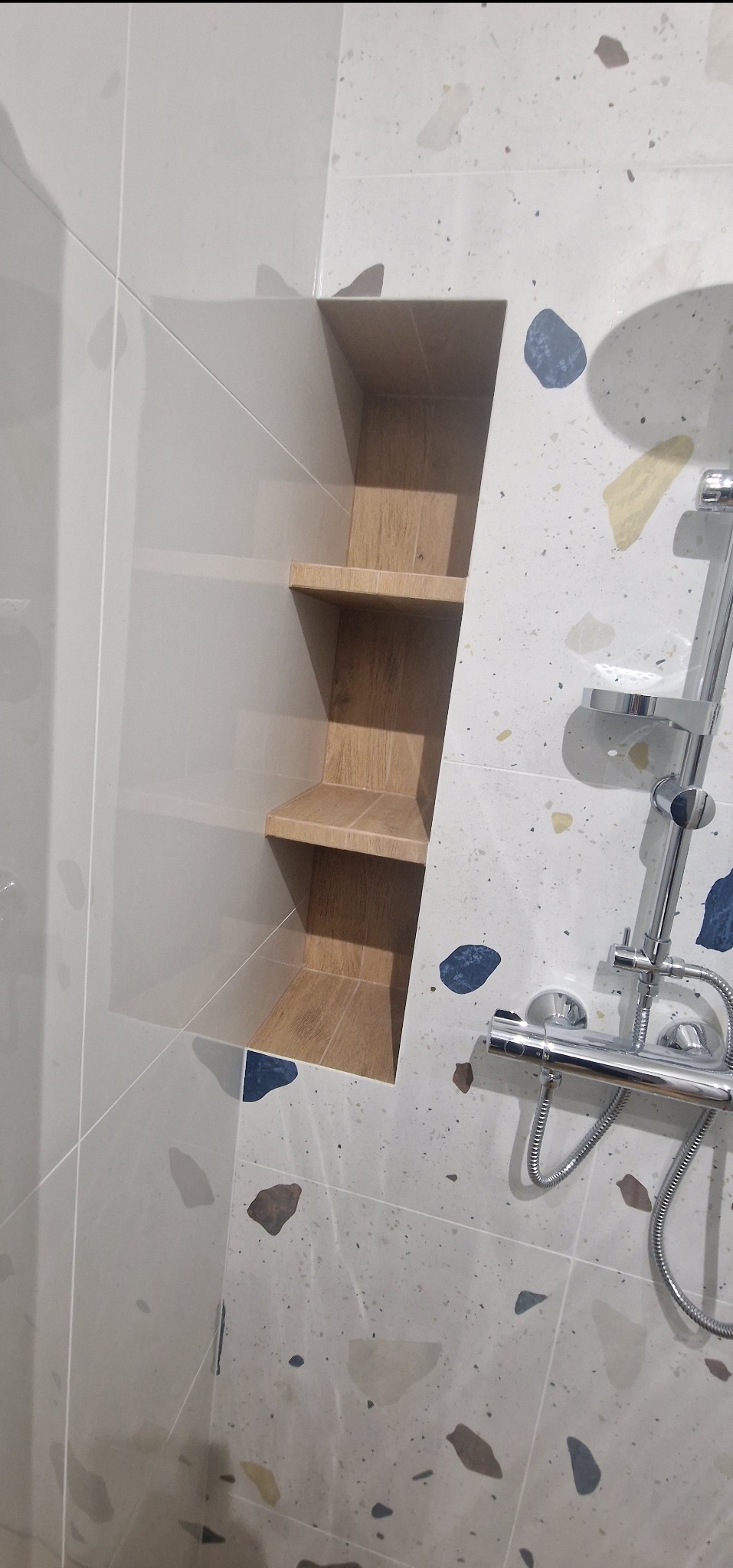 Ściana łazienki wyłożona płytkami w stylu terrazzo z wnęką z drewnianymi półkami i chromowaną baterią prysznicową.