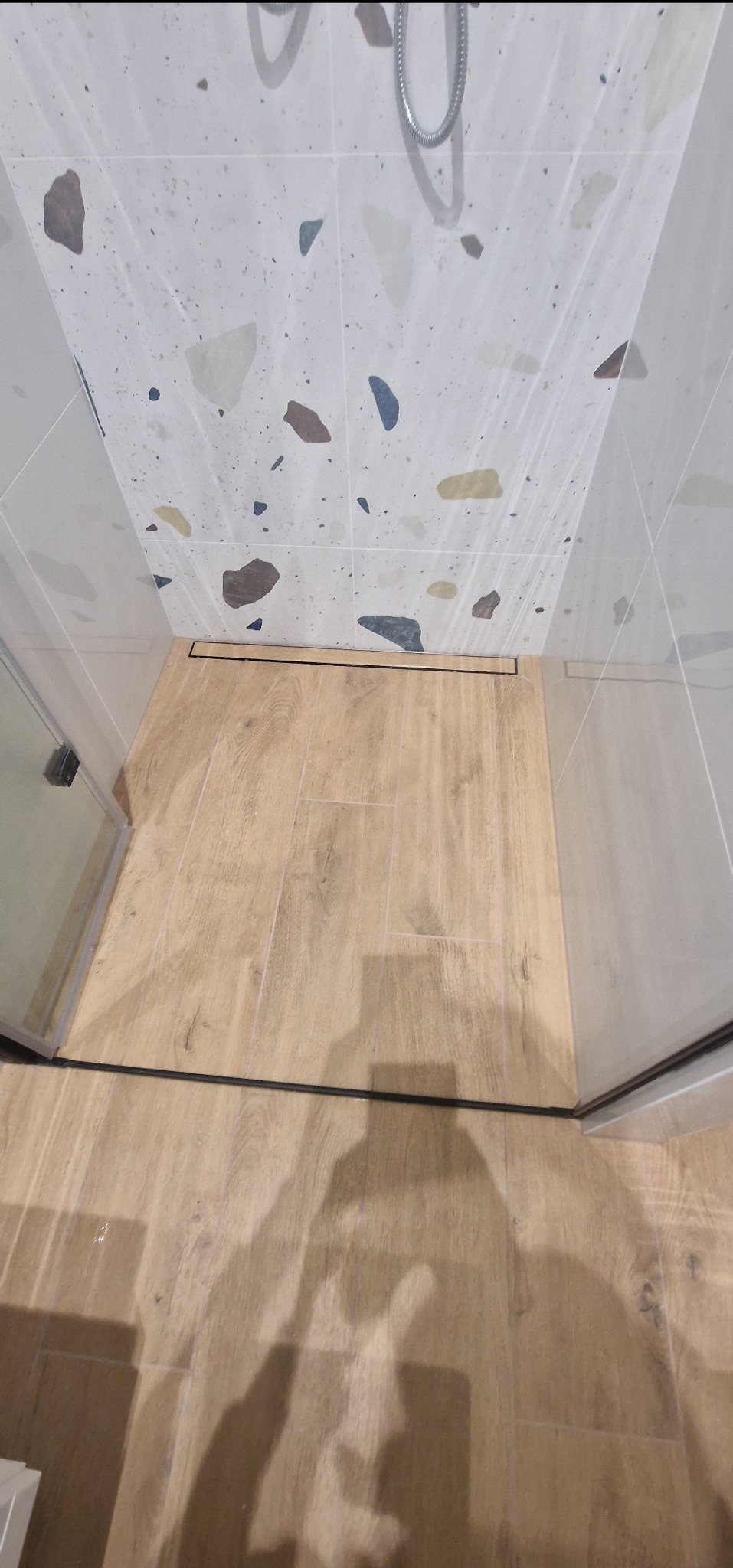 Wykończona kabina prysznicowa z odpływem liniowym, wyłożona płytkami imitującymi drewno na podłodze i mozaiką lastryko na ścianach.