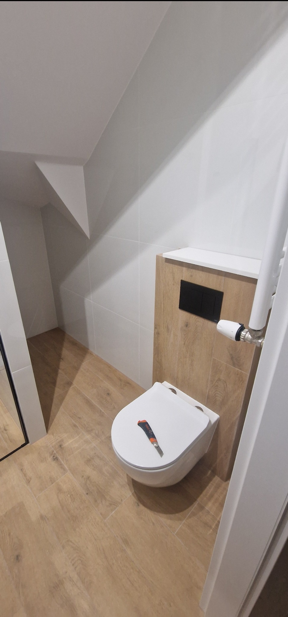 Świeżo wyremontowana łazienka na poddaszu z podwieszaną toaletą, na której leży nóż do tapet, drewnopodobna podłoga i białe płytki na ścianach.