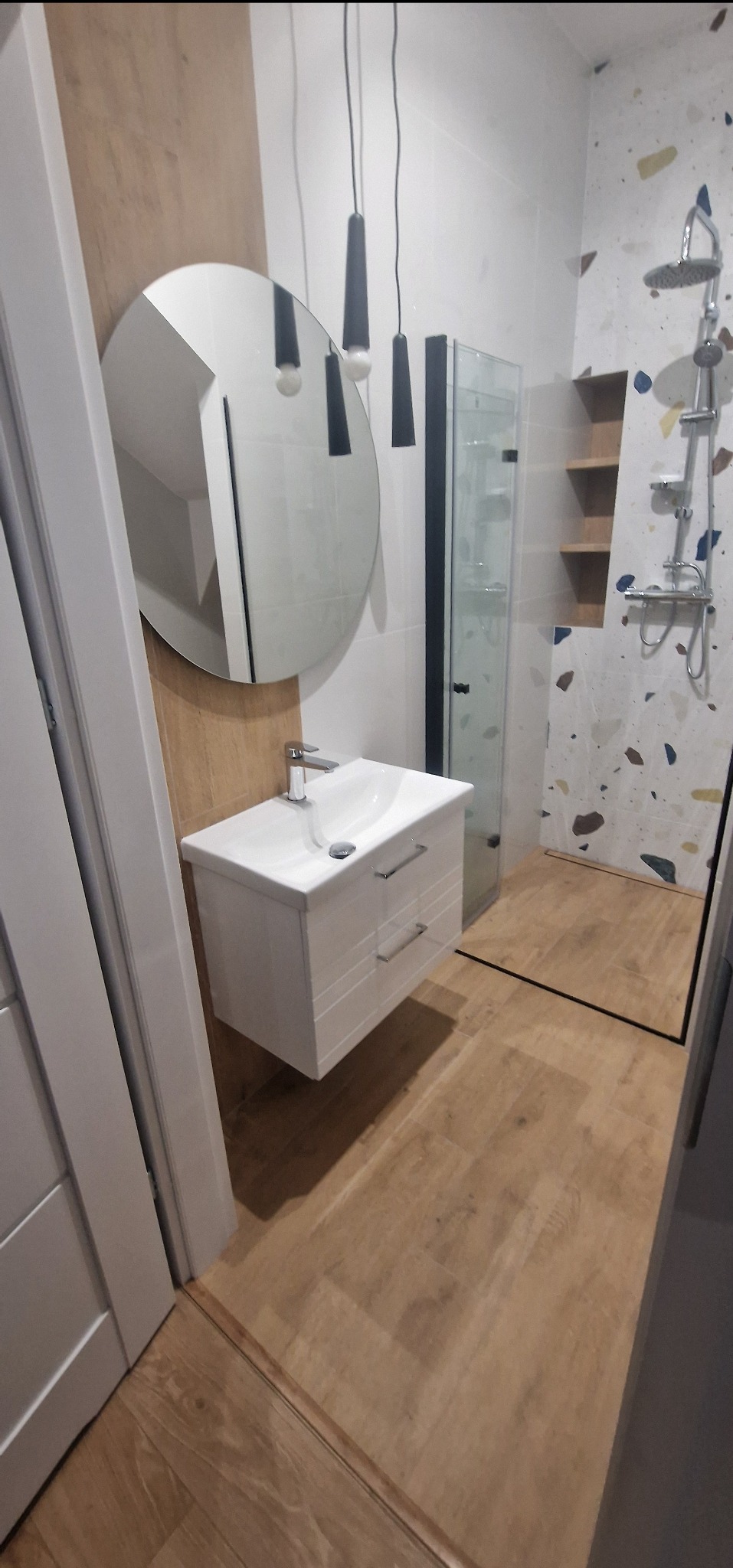 Nowoczesna łazienka z okrągłym lustrem nad białą szafką umywalkową, kabiną prysznicową z czarnymi ramami i ścianą wykończoną płytkami imitującymi lastryko.