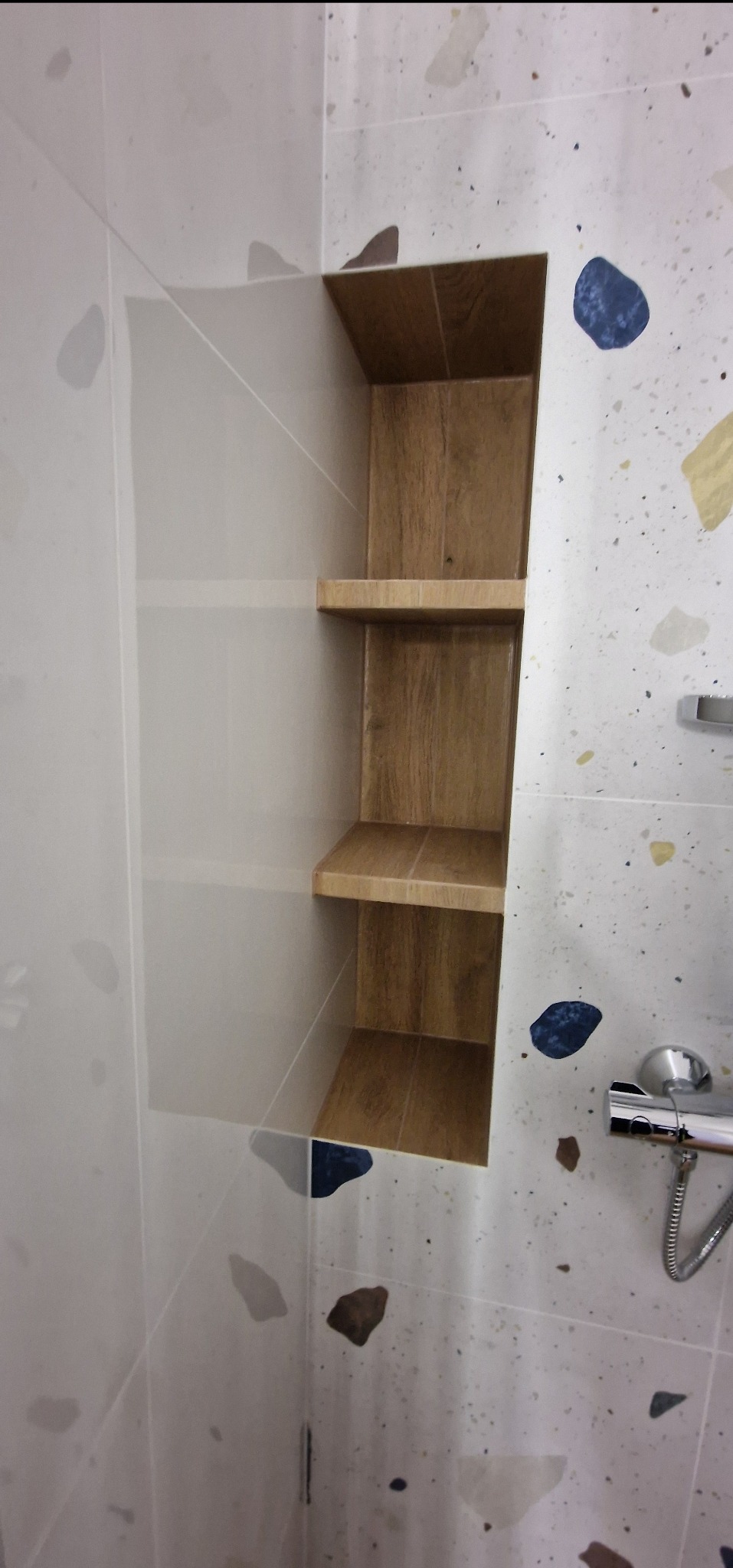 Wnęka z drewnopodobnymi półkami w wykończonej łazience z płytkami w stylu lastryko oraz chromowaną armaturą prysznicową.