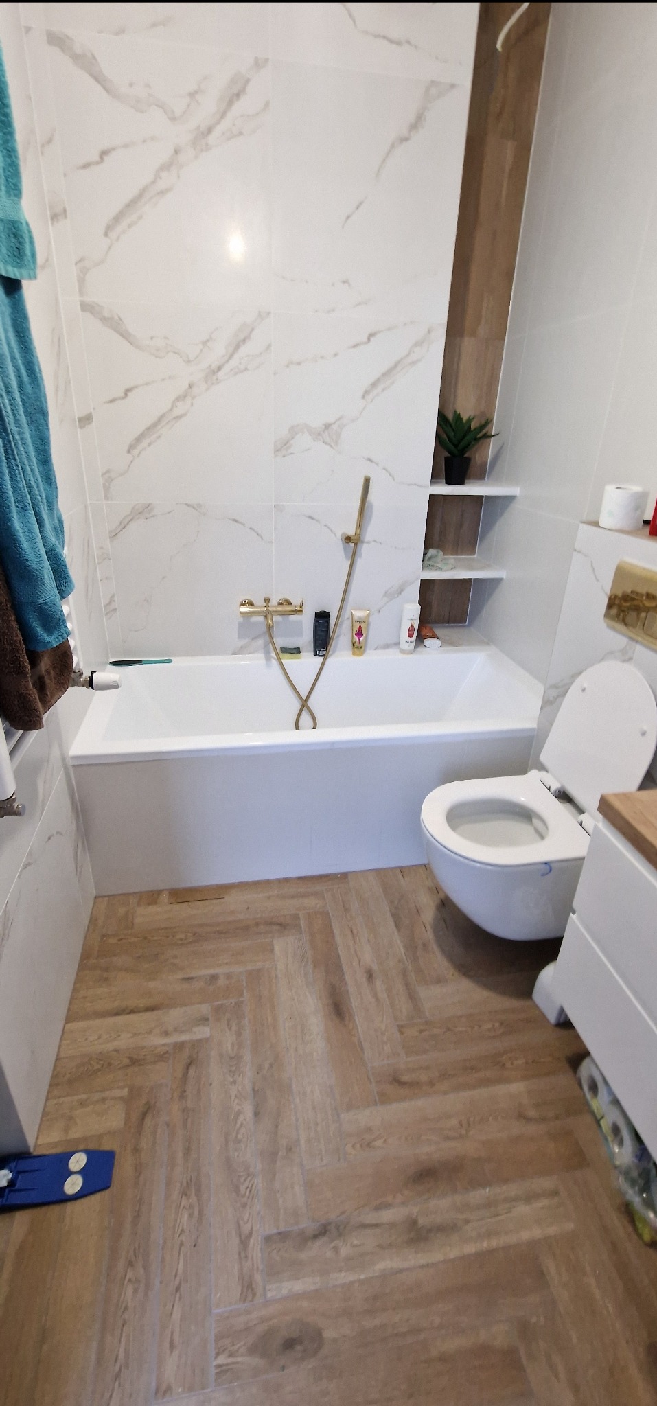 Biała łazienka z wanną, toaletą i drewnopodobną podłogą ułożoną w jodełkę, złota bateria wannowa i półki w zabudowie.