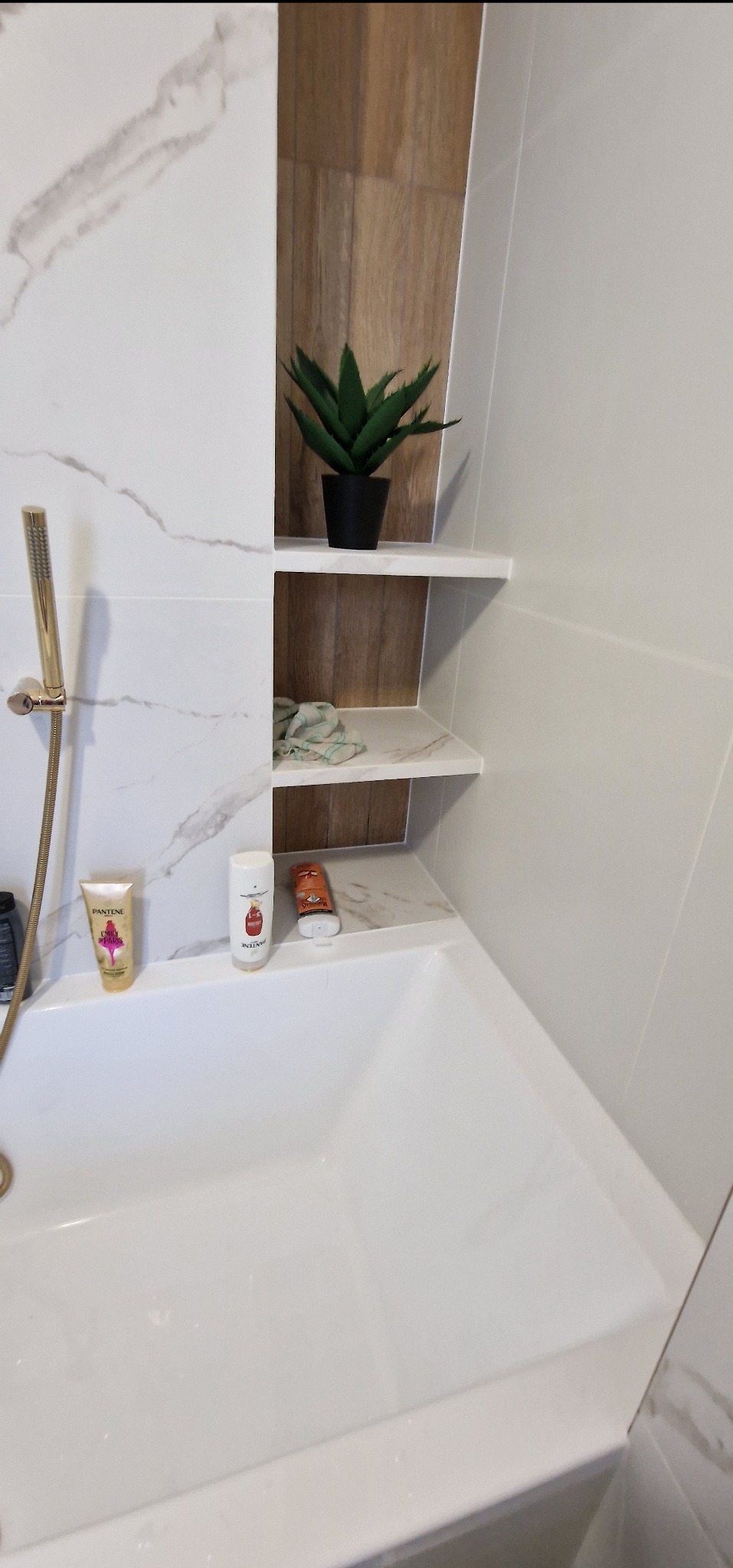 Nowoczesna łazienka z białą wanną, złotą słuchawką prysznicową, marmurkowymi płytkami i drewnianą wnęką z półkami, na których stoi sztuczna roślina i kosmetyki.