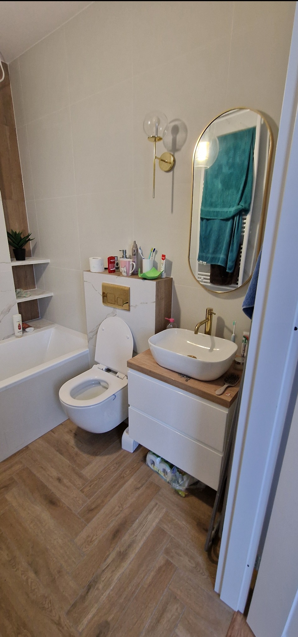 Nowoczesna łazienka z toaletą, umywalką nablatową na szafce z drewnianym blatem, złotą armaturą, lustrem w złotej ramie i podłogą w jodełkę.