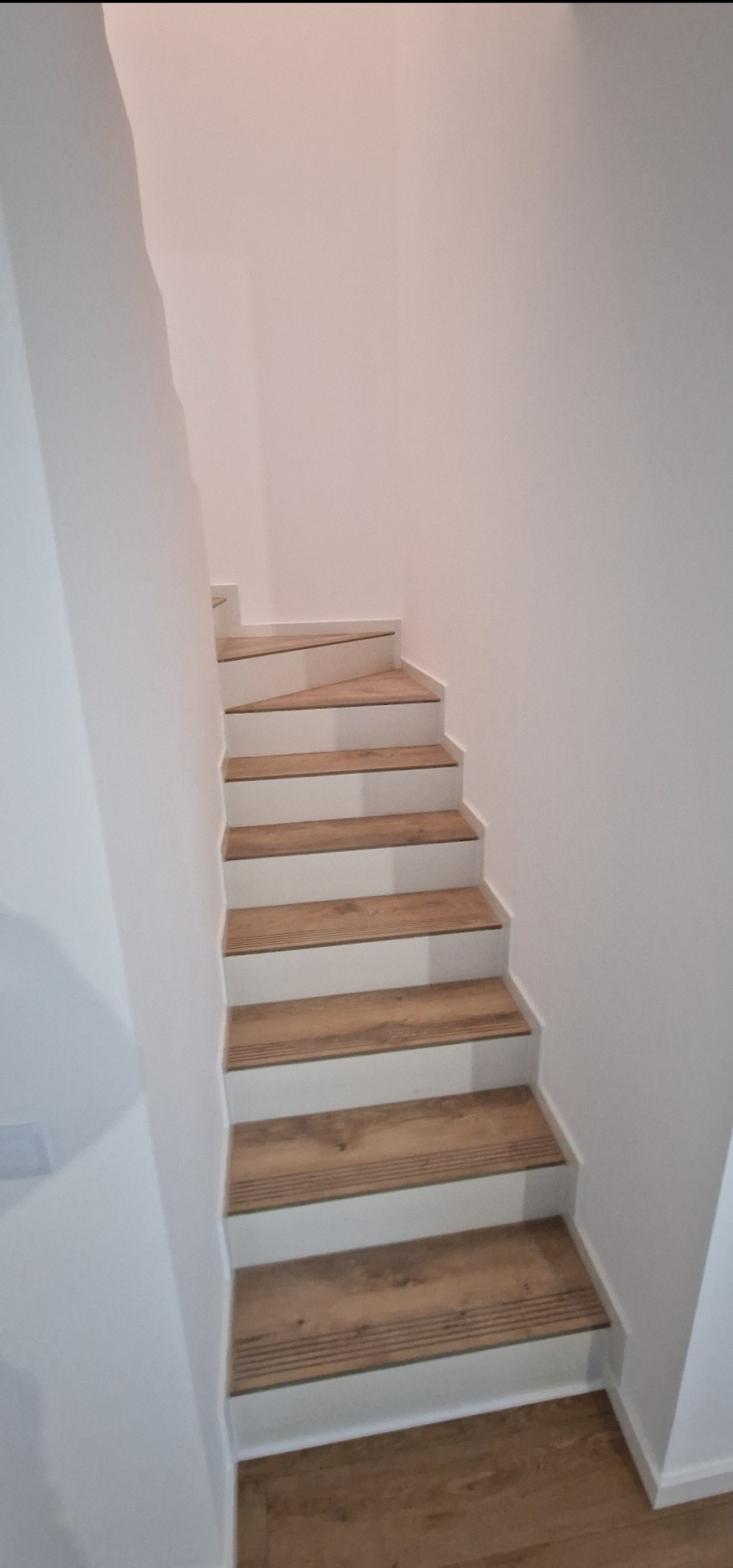 Bieg schodów w jasnej klatce schodowej, wykończony panelami imitującymi drewno, z białymi podstopnicami, widziany z dołu.