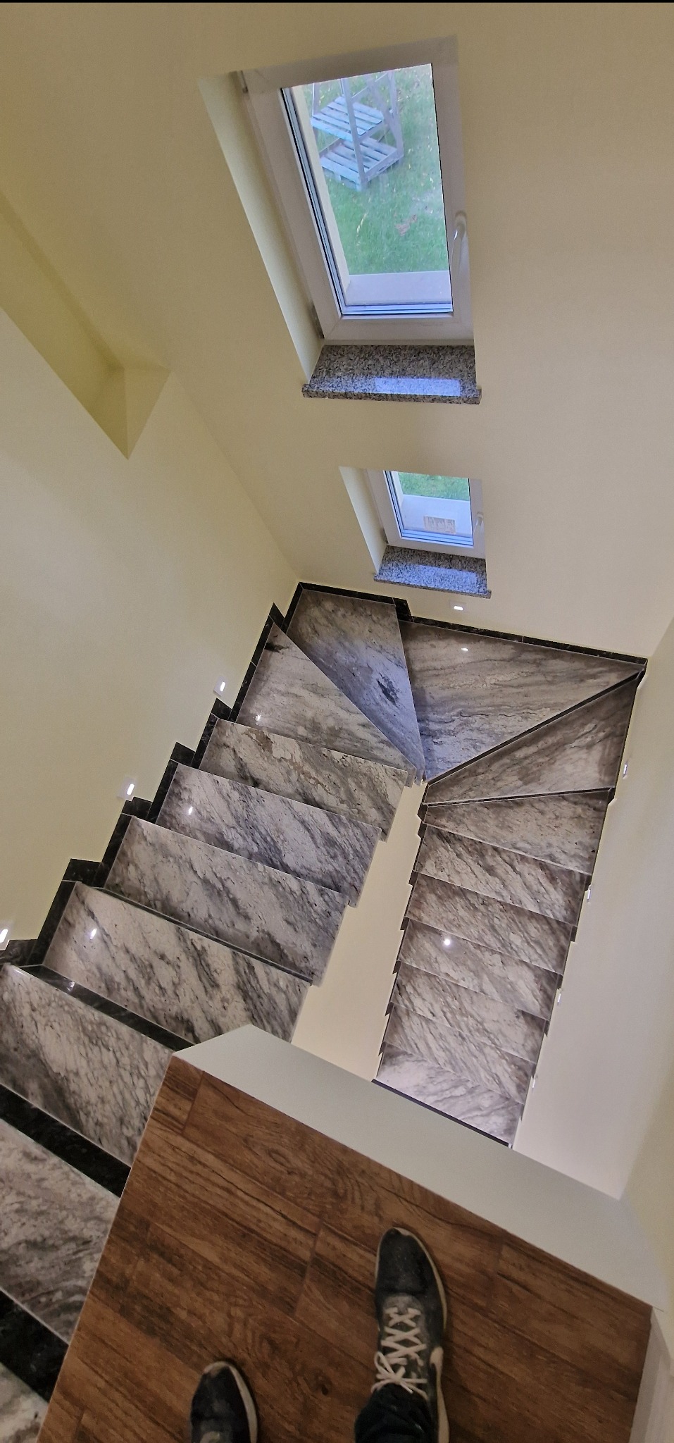 Schody wykończone marmurem z podświetleniem LED, widok z góry, z fragmentem drewnianej podłogi i butami. Dwa okna dachowe w tle.