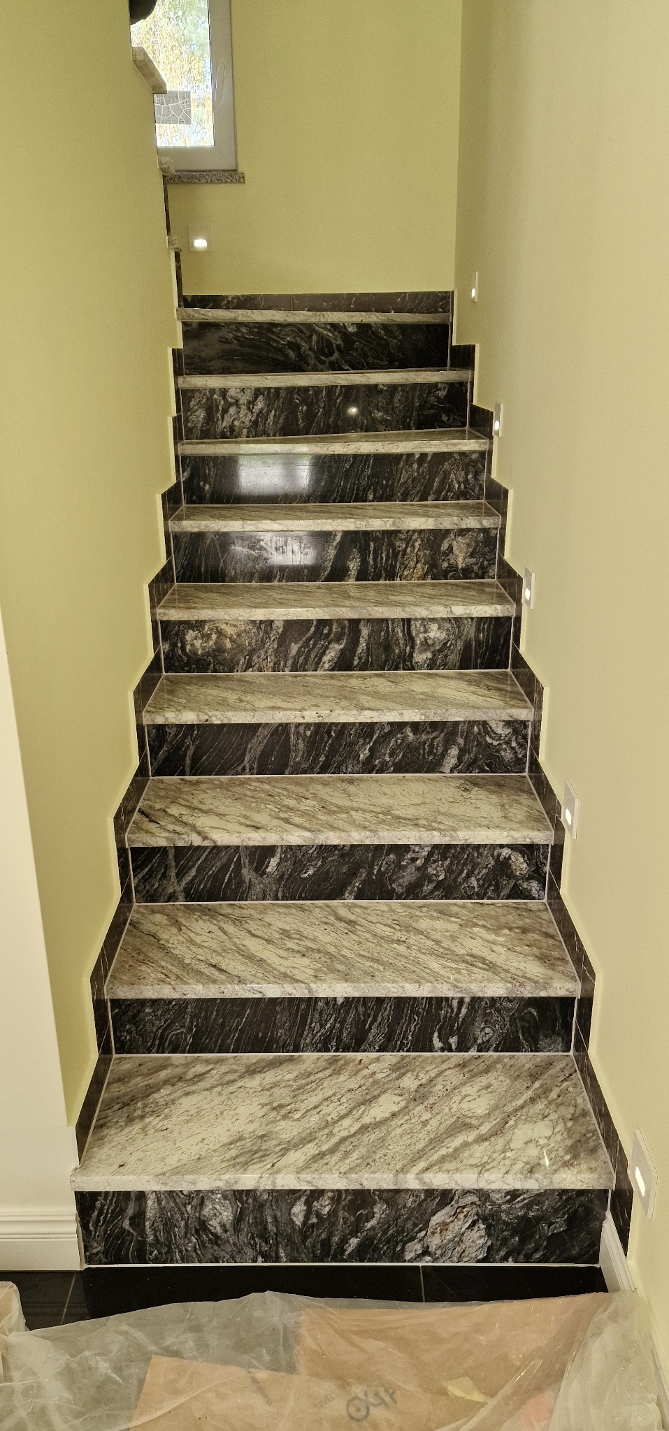 Schody wyłożone marmurem w odcieniach szarości i czerni, z podświetleniem LED na ścianie, widoczne zabezpieczenia podłogi.