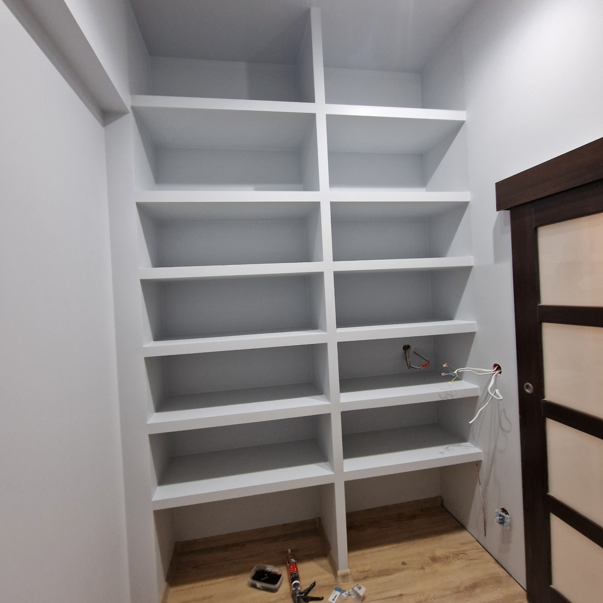 Wnęka z białymi półkami w trakcie wykańczania, widoczne wystające przewody elektryczne i narzędzia na podłodze, obok drzwi z ciemną ramą.