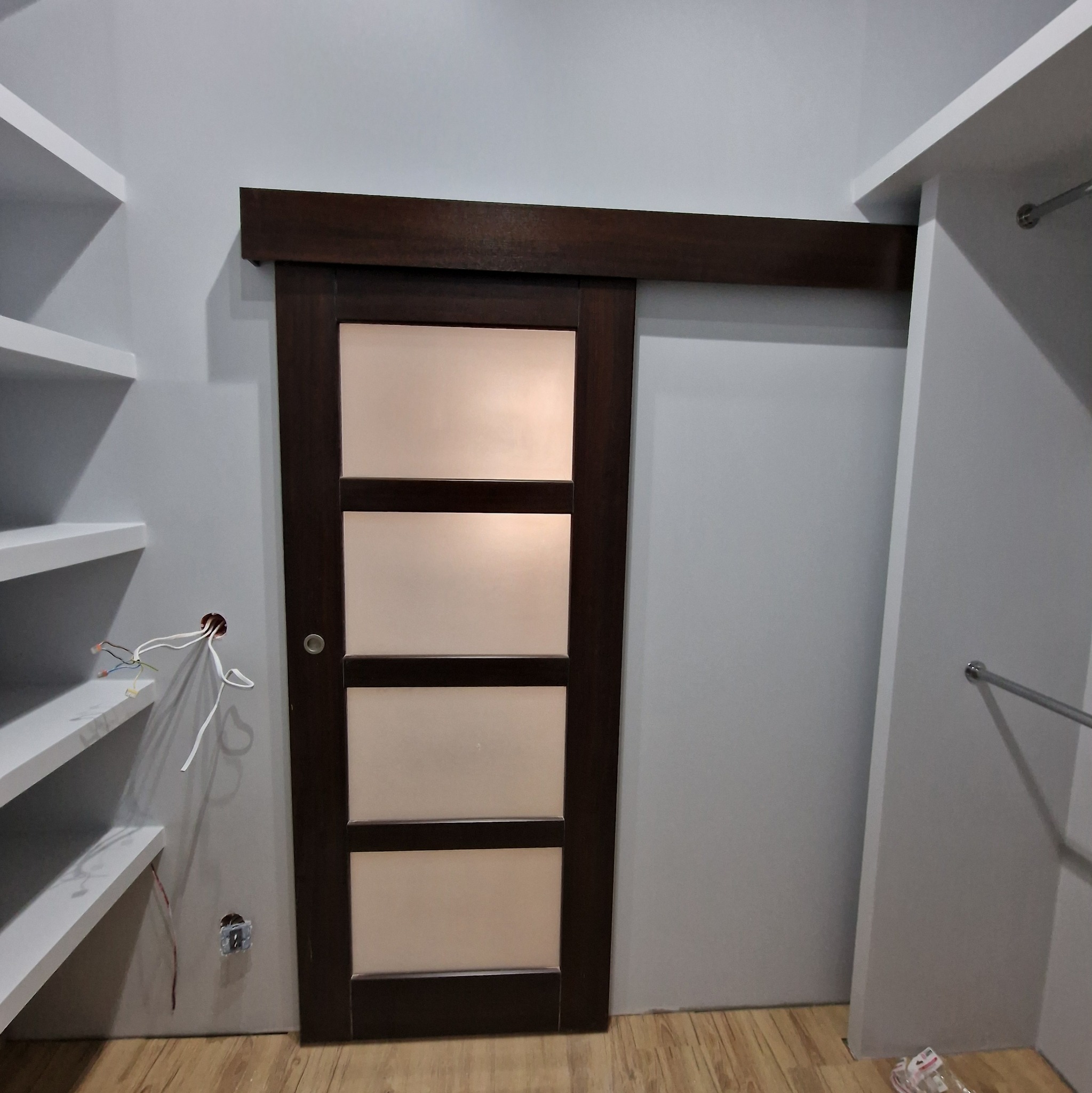 Brązowe drzwi przesuwne z czterema matowymi szybami zamontowane w garderobie z białymi półkami, widoczne wystające przewody elektryczne.