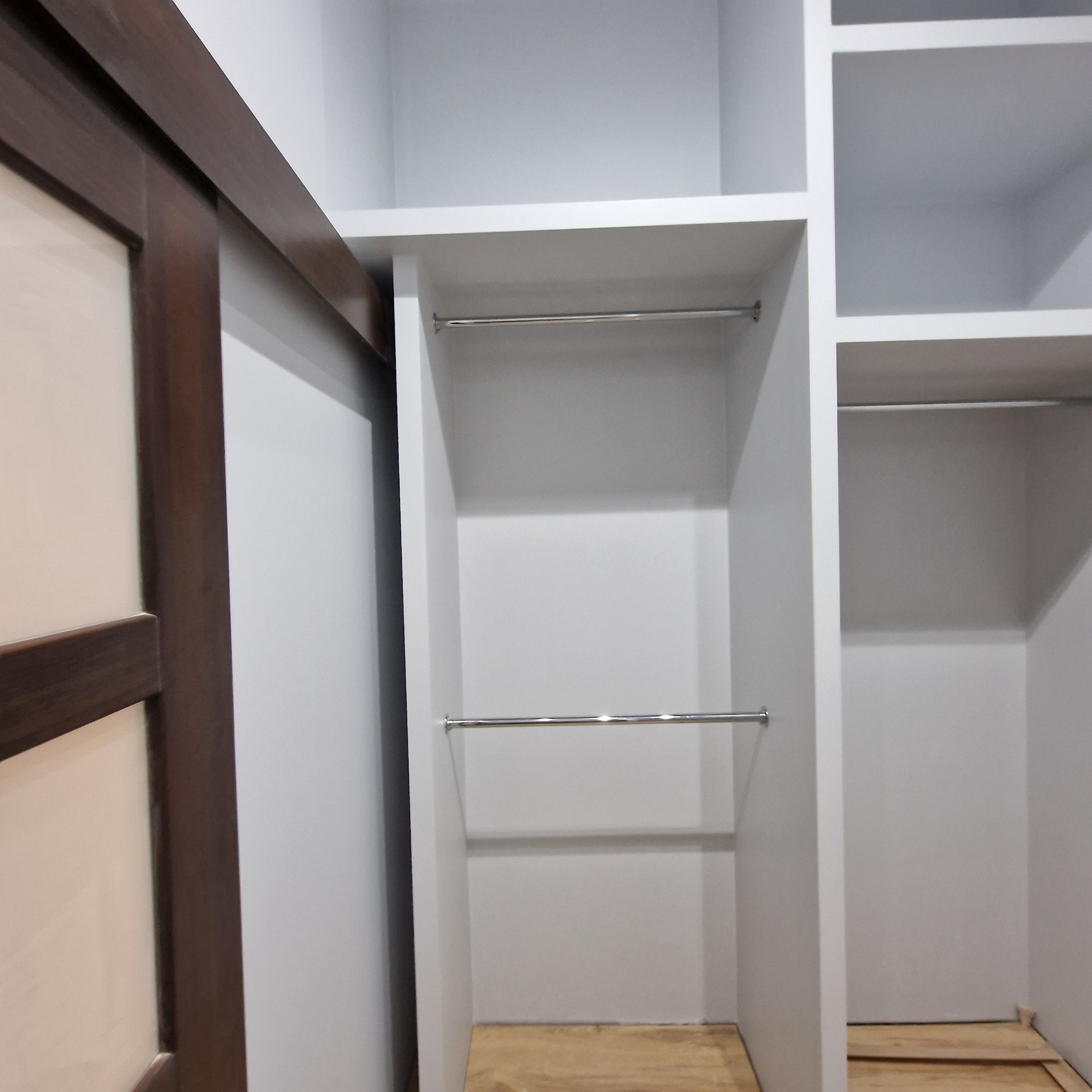 Biała, modułowa garderoba z przesuwnymi drzwiami z ciemnego drewna, wyposażona w metalowe drążki na ubrania, na podłodze widoczne deski.