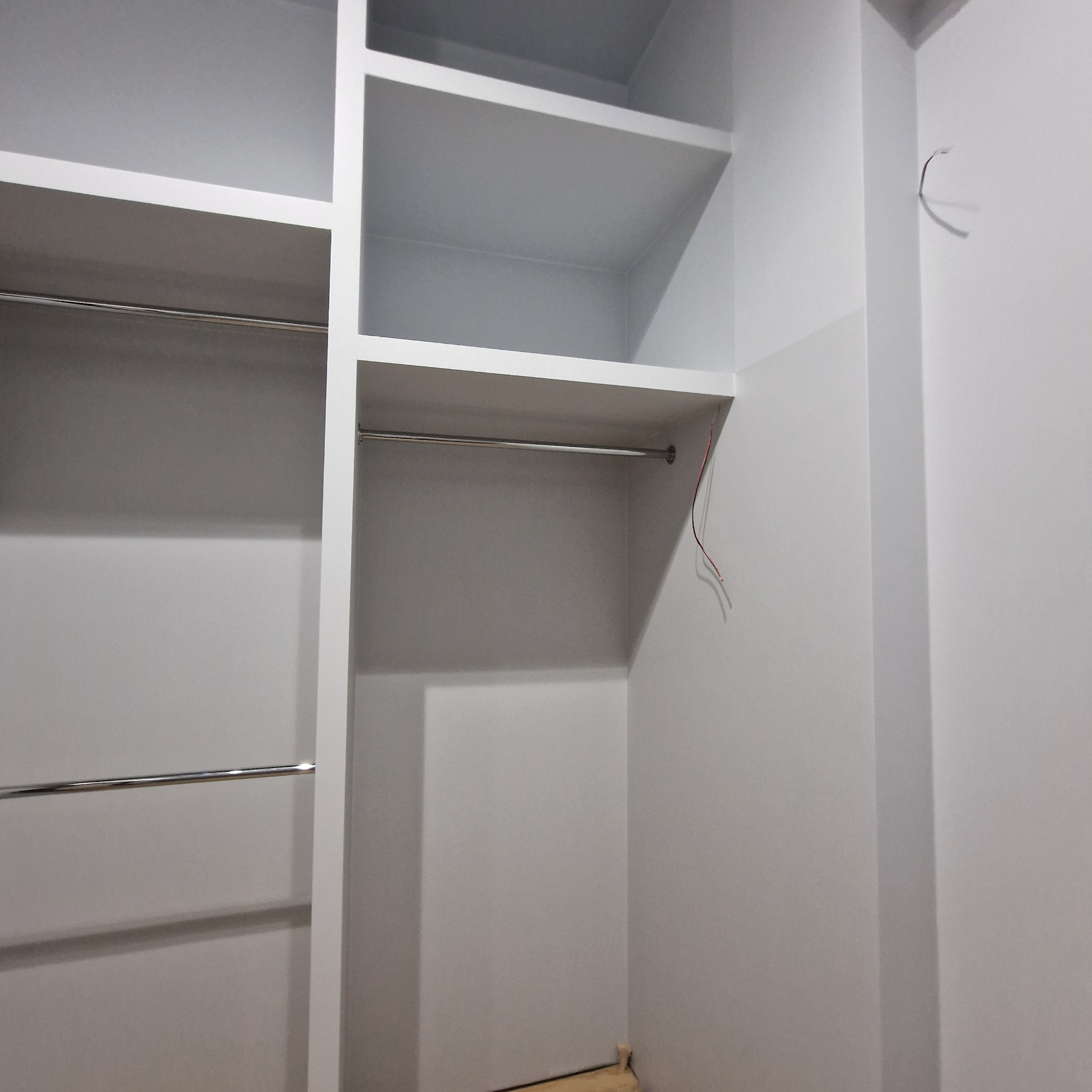 Wnętrze szafy wnękowej z półkami i drążkami na ubrania, świeżo pomalowane ściany na szaro, widoczne wystające kable elektryczne.
