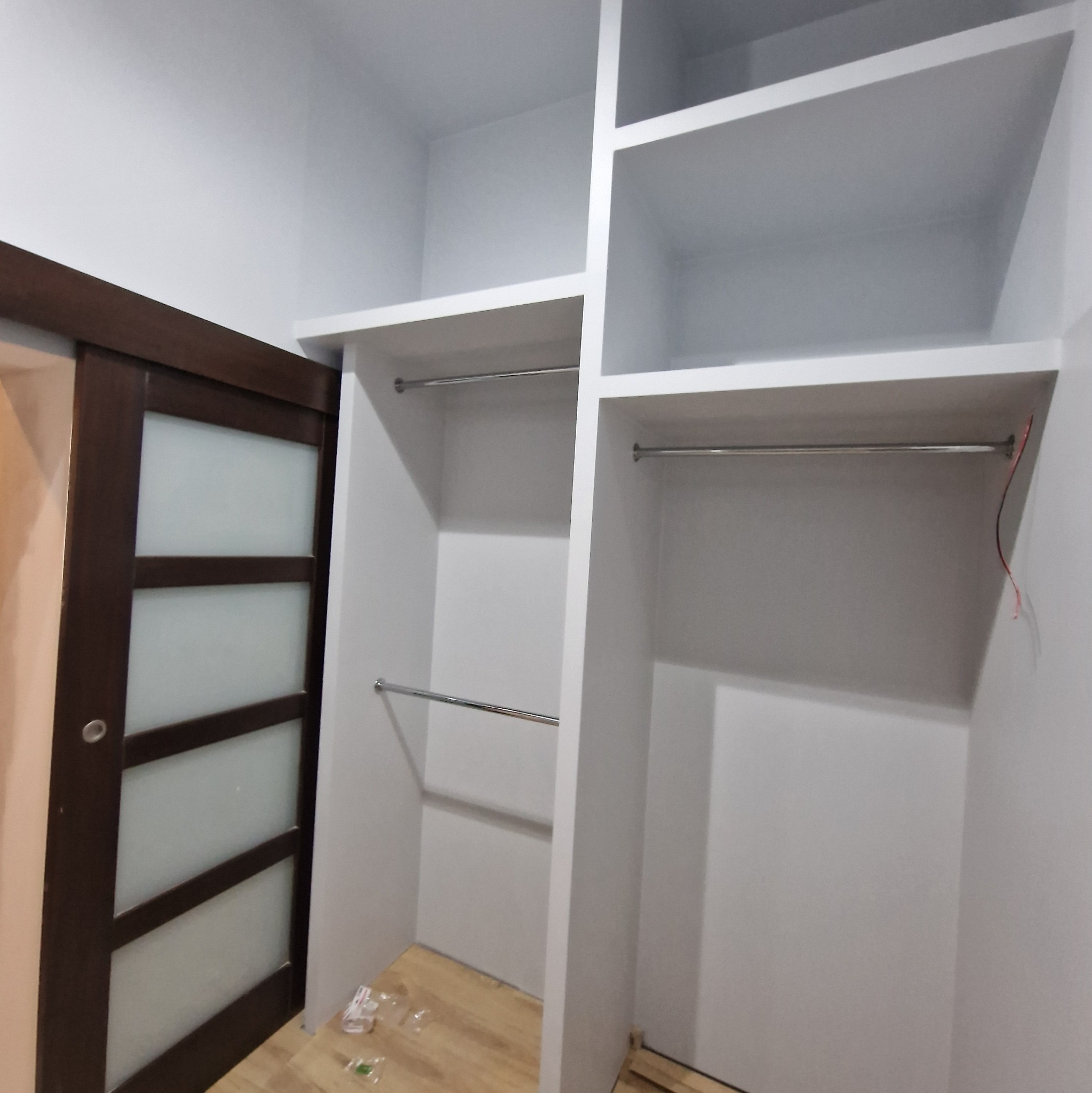 Wnętrze garderoby z białymi półkami i drążkami na ubrania, z częściowo widocznymi przesuwnymi drzwiami z ciemnego drewna i matowego szkła, na podłodze widoczne drobne śmieci po montażu.