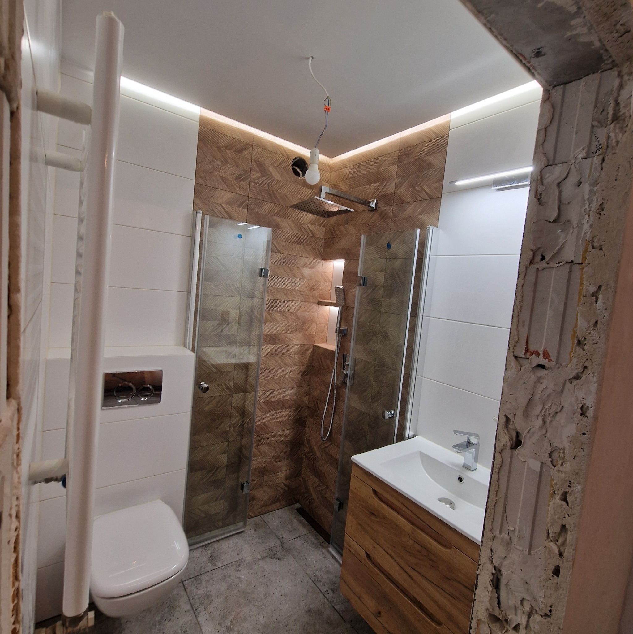 Nowoczesna łazienka z kabiną prysznicową typu walk-in, drewnianą szafką pod umywalką i wiszącą toaletą, widoczny surowy fragment ściany przed wykończeniem.