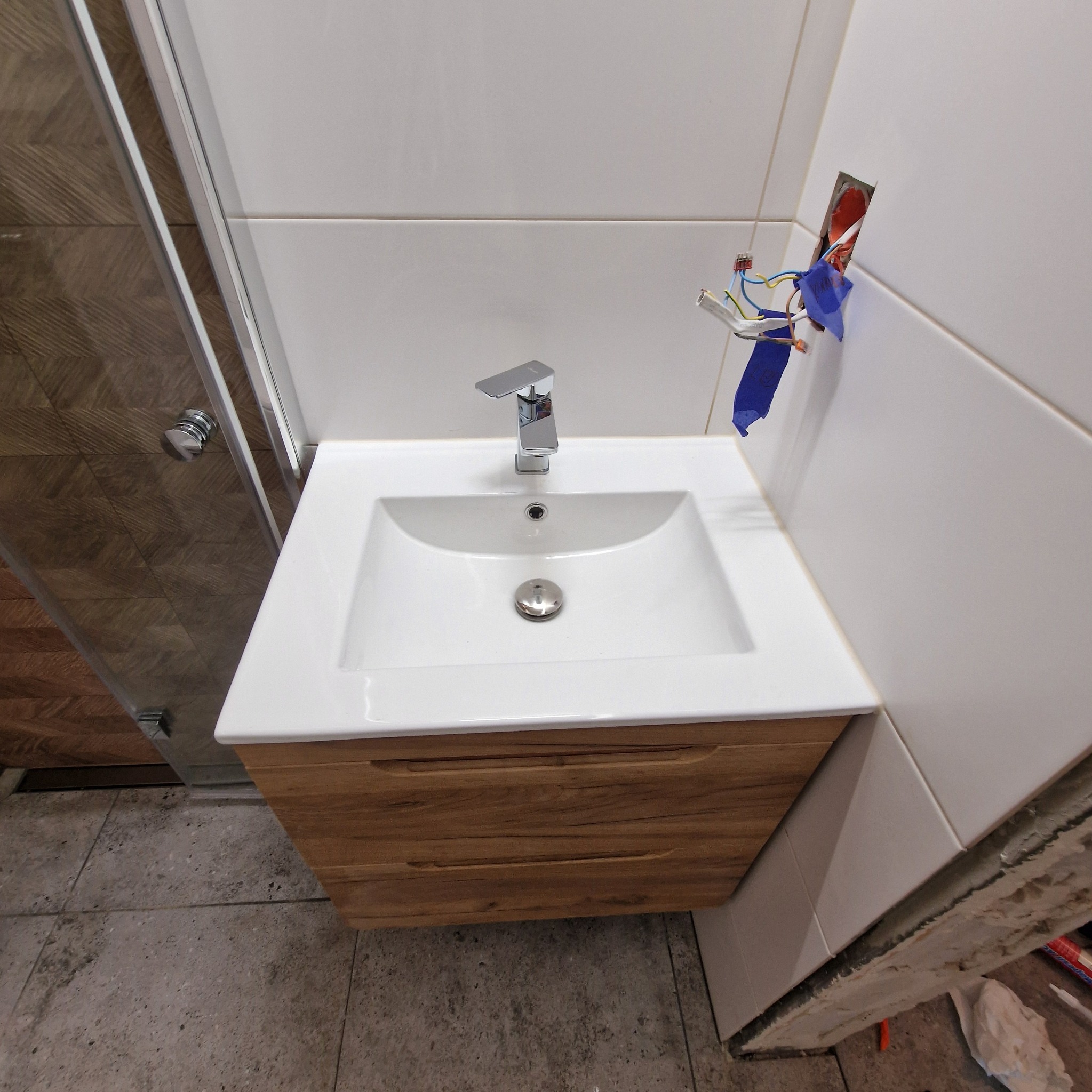 Biała umywalka z drewnianą szafką w łazience w trakcie remontu, widoczne niezabezpieczone przewody elektryczne i fragment kabiny prysznicowej.