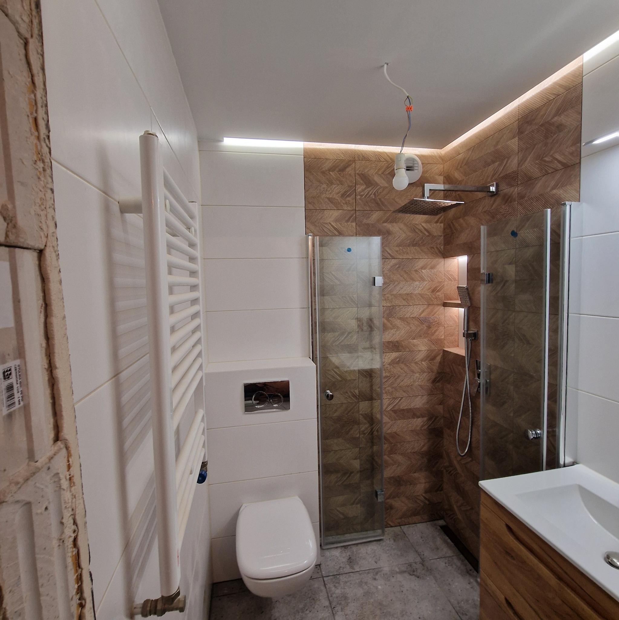 Nowoczesna łazienka z drewnopodobnymi płytkami ułożonymi w jodełkę na ścianie prysznica, białe płytki na pozostałych ścianach, podwieszana toaleta, grzejnik drabinkowy i drewniana szafka...