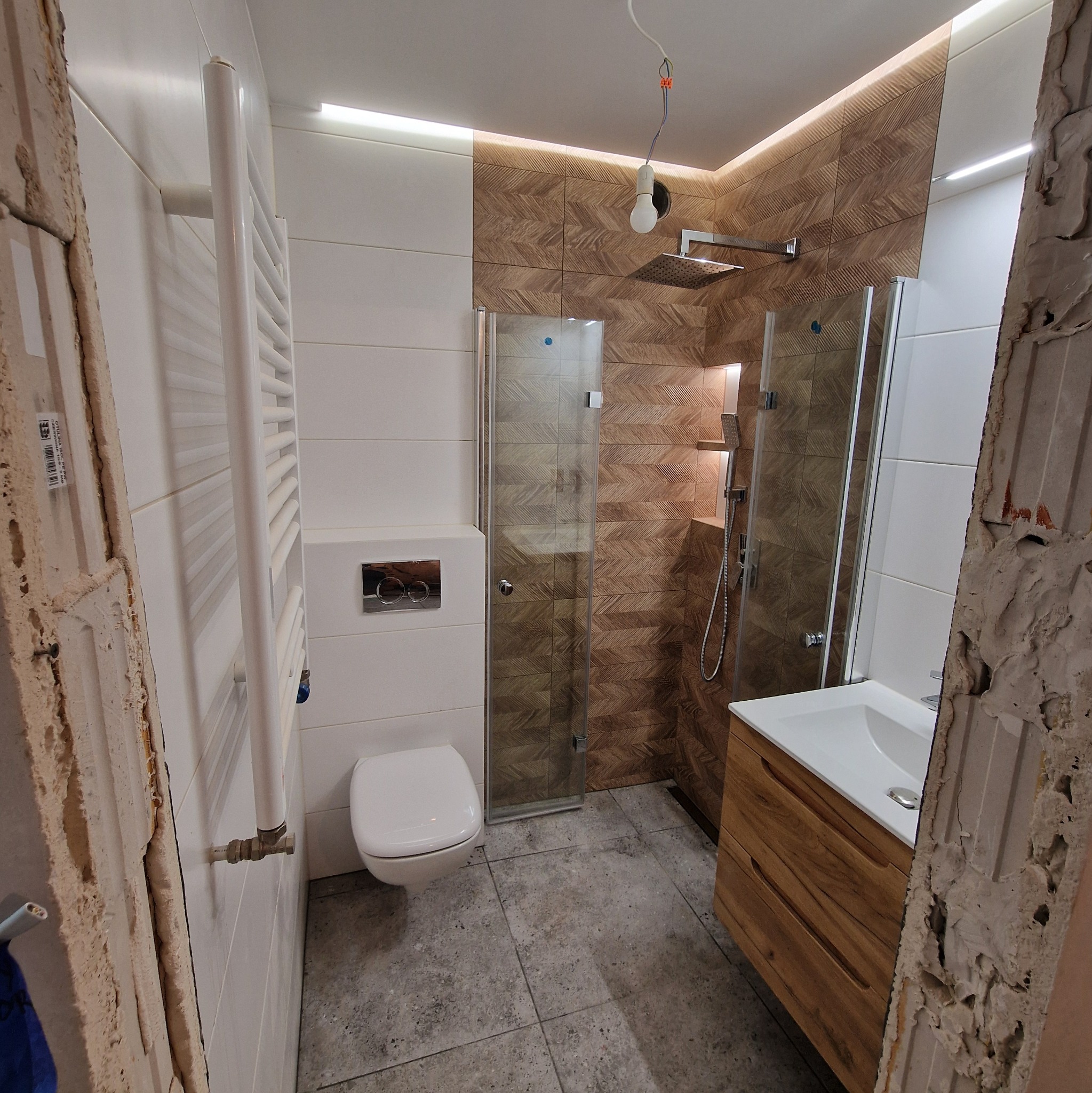 Częściowo wykończona łazienka z widocznymi fragmentami ściany w stanie surowym, nowoczesna kabina prysznicowa z deszczownicą, umywalka z szafką z drewnopodobnym wykończeniem, biała toaleta wisząca...