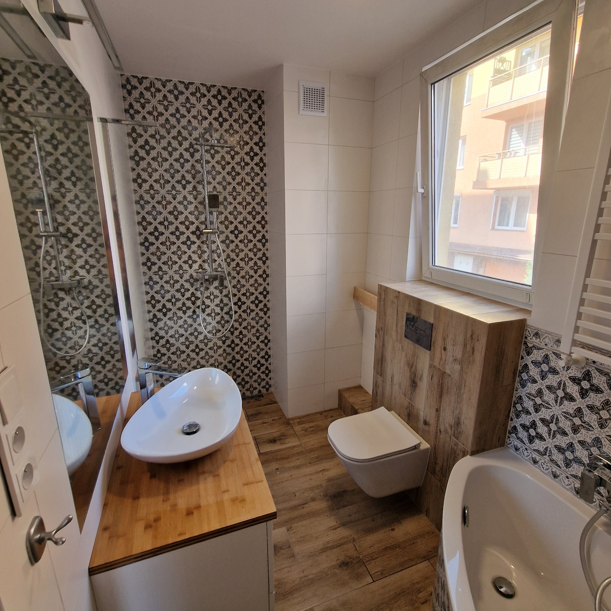 Nowoczesna łazienka z umywalką nablatową na drewnianym blacie, wiszącą toaletą z obudową imitującą drewno, płytkami w geometryczne wzory i widokiem z okna na budynek mieszkalny.