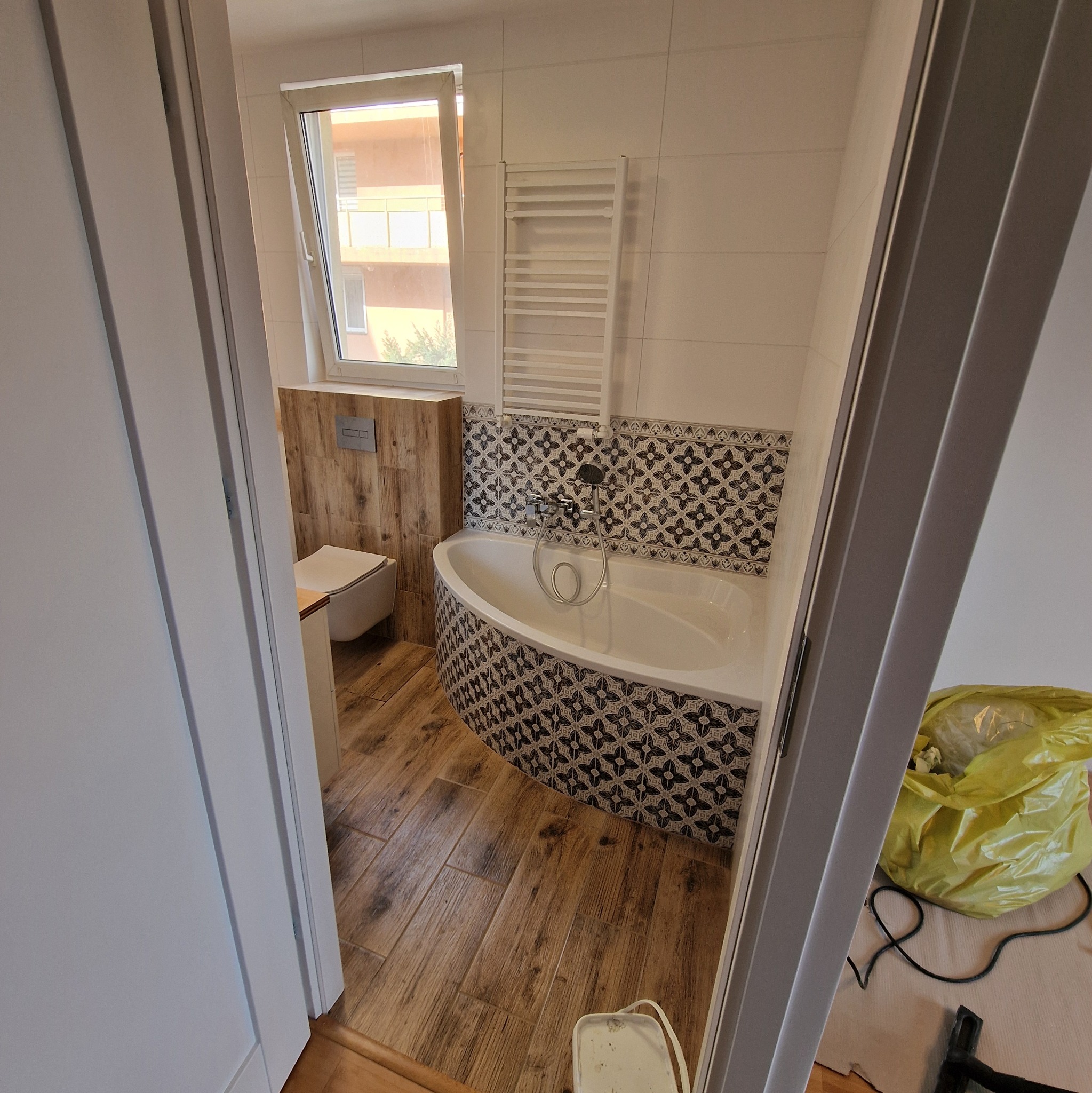 Wykończona łazienka z drewnopodobną podłogą, obudowaną wanną z dekoracyjnymi kaflami, grzejnikiem drabinkowym i drewnianą zabudową toalety.